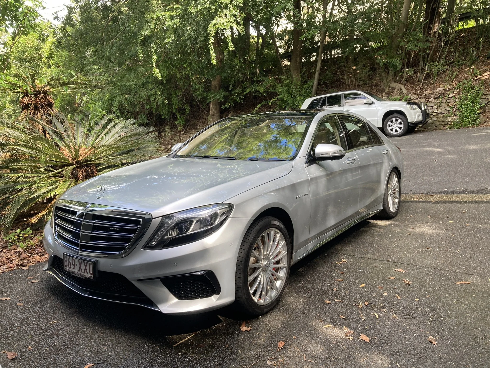 A silver Mercedes-Benz sedan parked on a driveway with a white SUV in the background, surrounded by lush green trees and bushes.