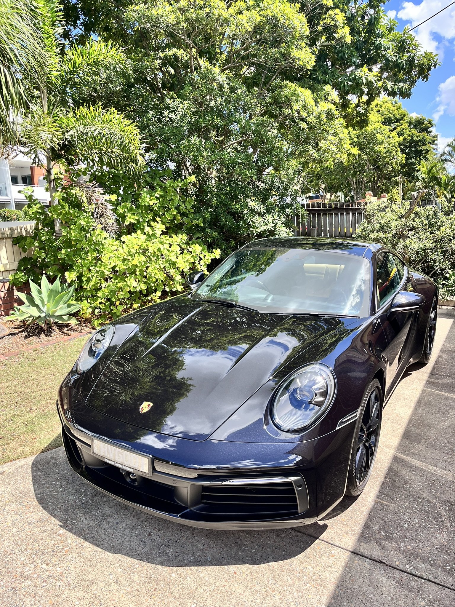Black Porsche sports car parked on driveway with green trees and bushes in the background under a blue sky with clouds.