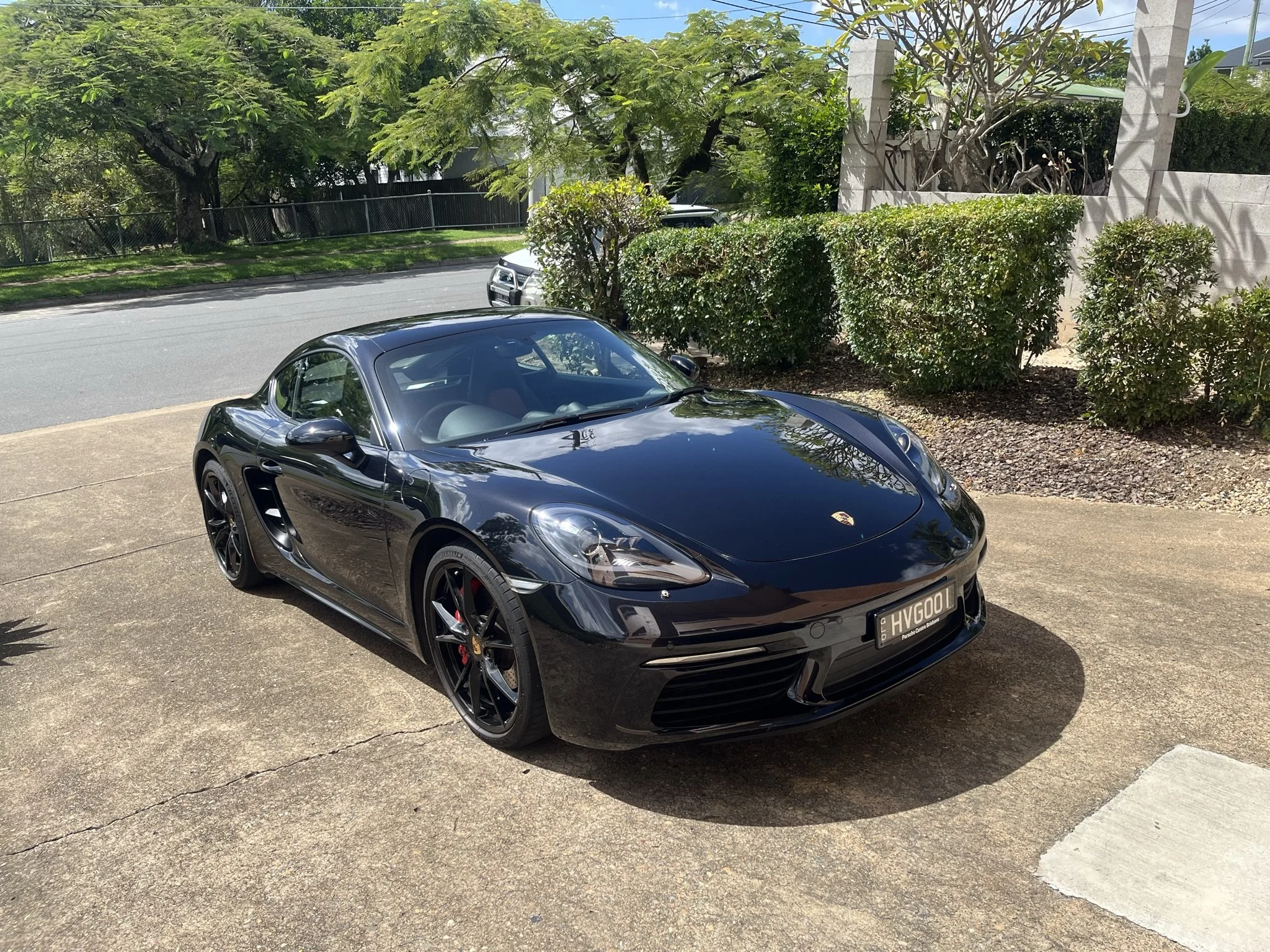A black Porsche sports car parked on a driveway with greenery and trees in the background.