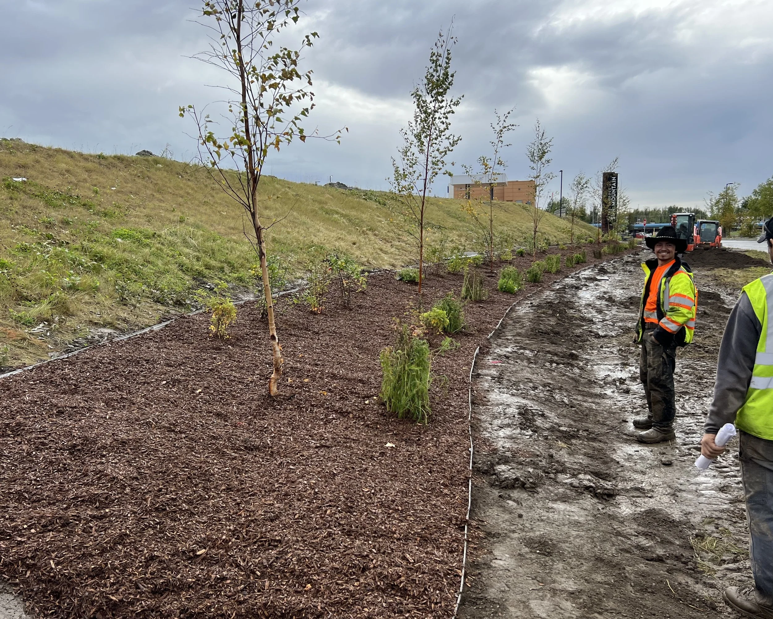 Two workers in safety vests and hats standing on a muddy construction site near a landscaped area with young trees and shrubs, with cloudy sky overhead.