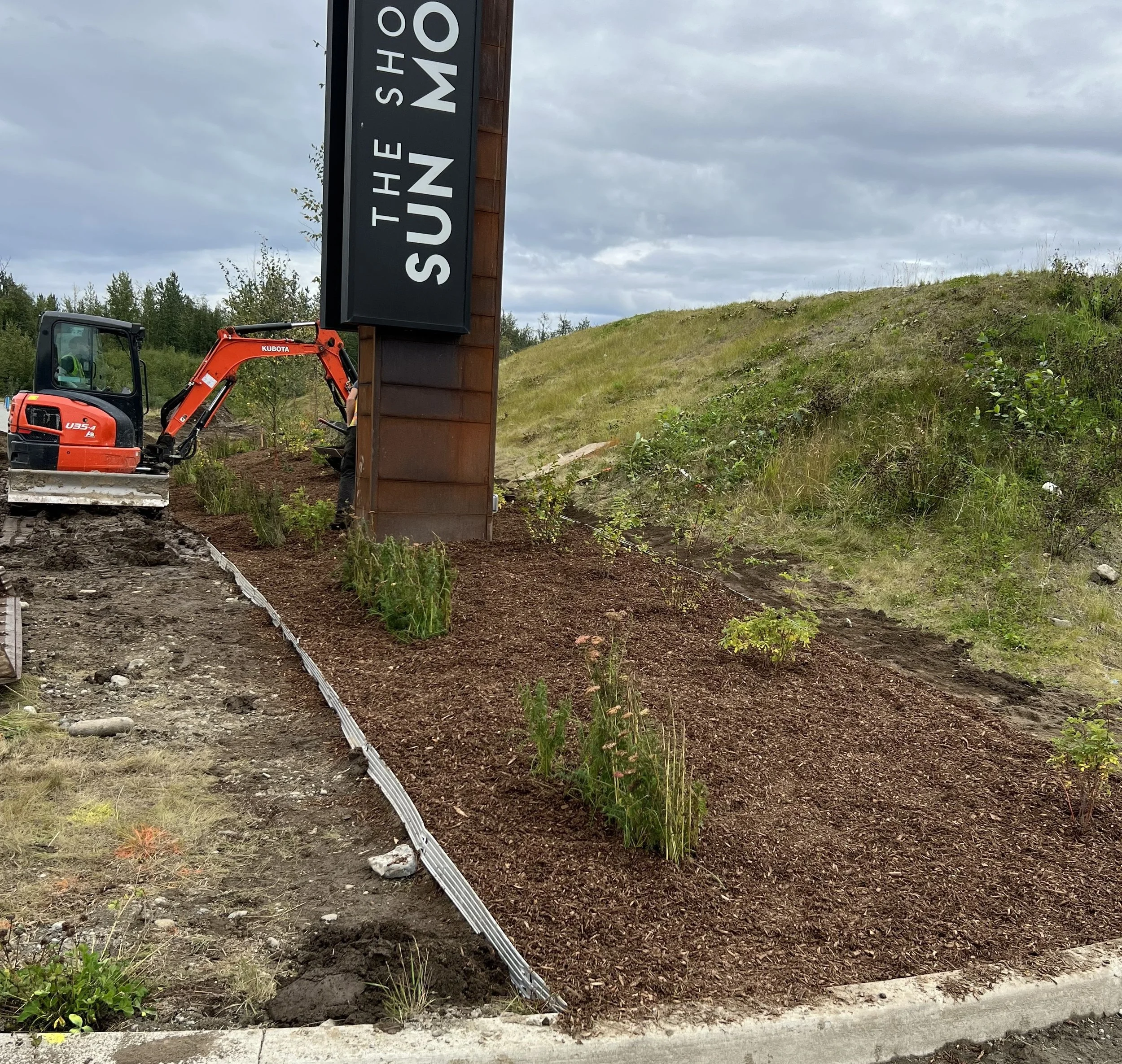 A small orange and black mini excavator working on landscaping around a sign that reads 'The Sun Mo'. The area has freshly planted bushes and mulch, with a grassy hillside in the background under cloudy skies.
