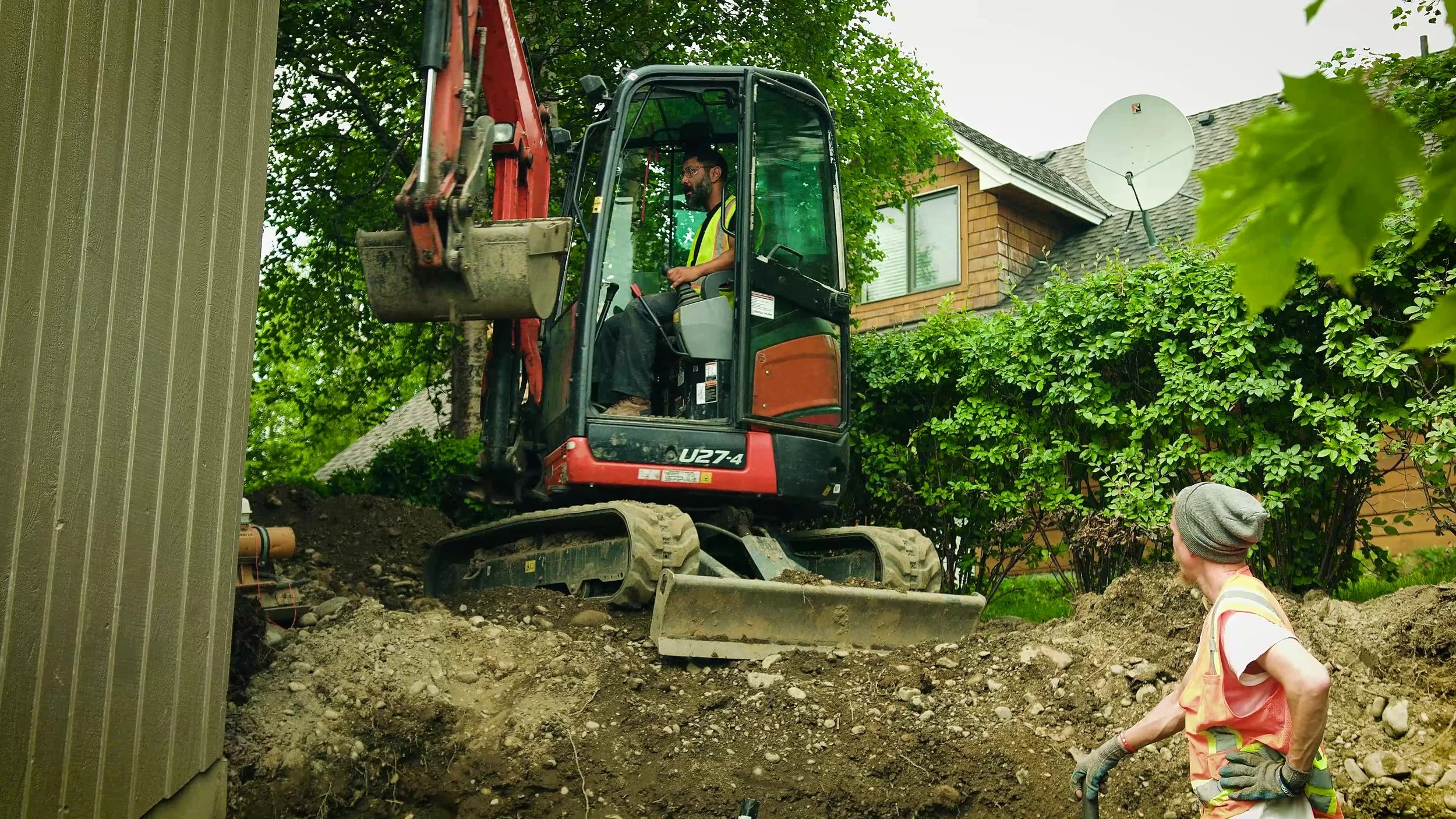 Walker Landscaper expert heavy machine operatoroperating a small red excavator near a house in Wasilla with greenery in the background.