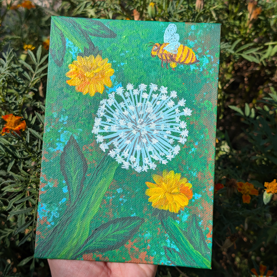 Hand holding a small canvas painting of a dandelion with two yellow flowers and a bee against a green background, outdoors among green foliage.