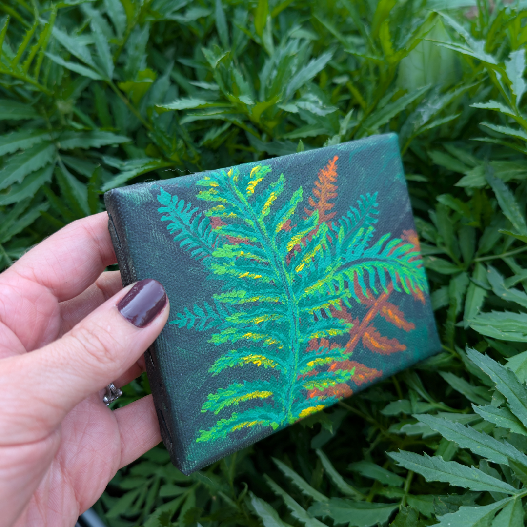 Hand holding a small canvas with a painted green fern leaf, surrounded by lush green foliage.