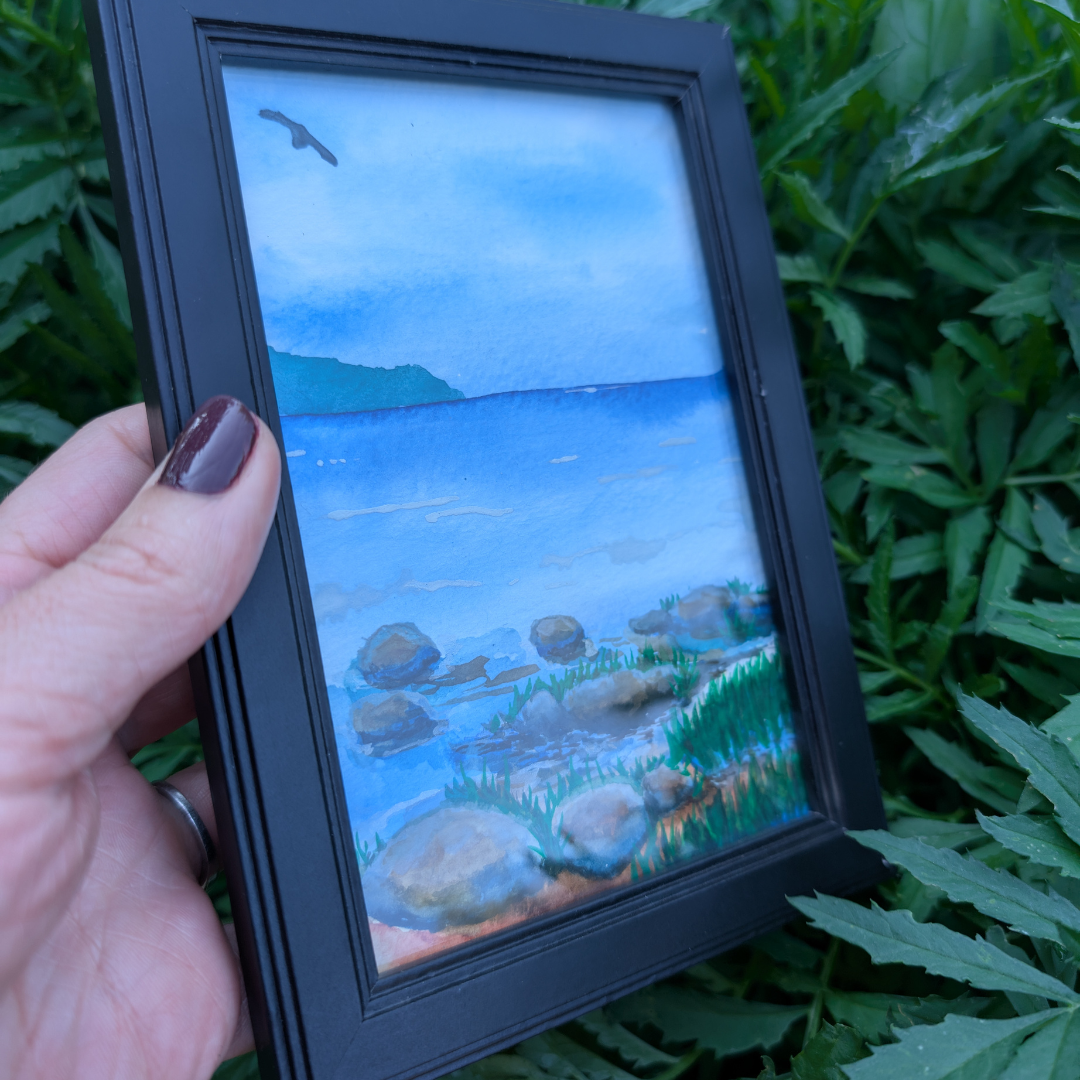 A person holding a framed watercolor painting of a lakeside scene with rocks, grass, and a distant mountain, reflecting a cloudy sky and a bird flying overhead.