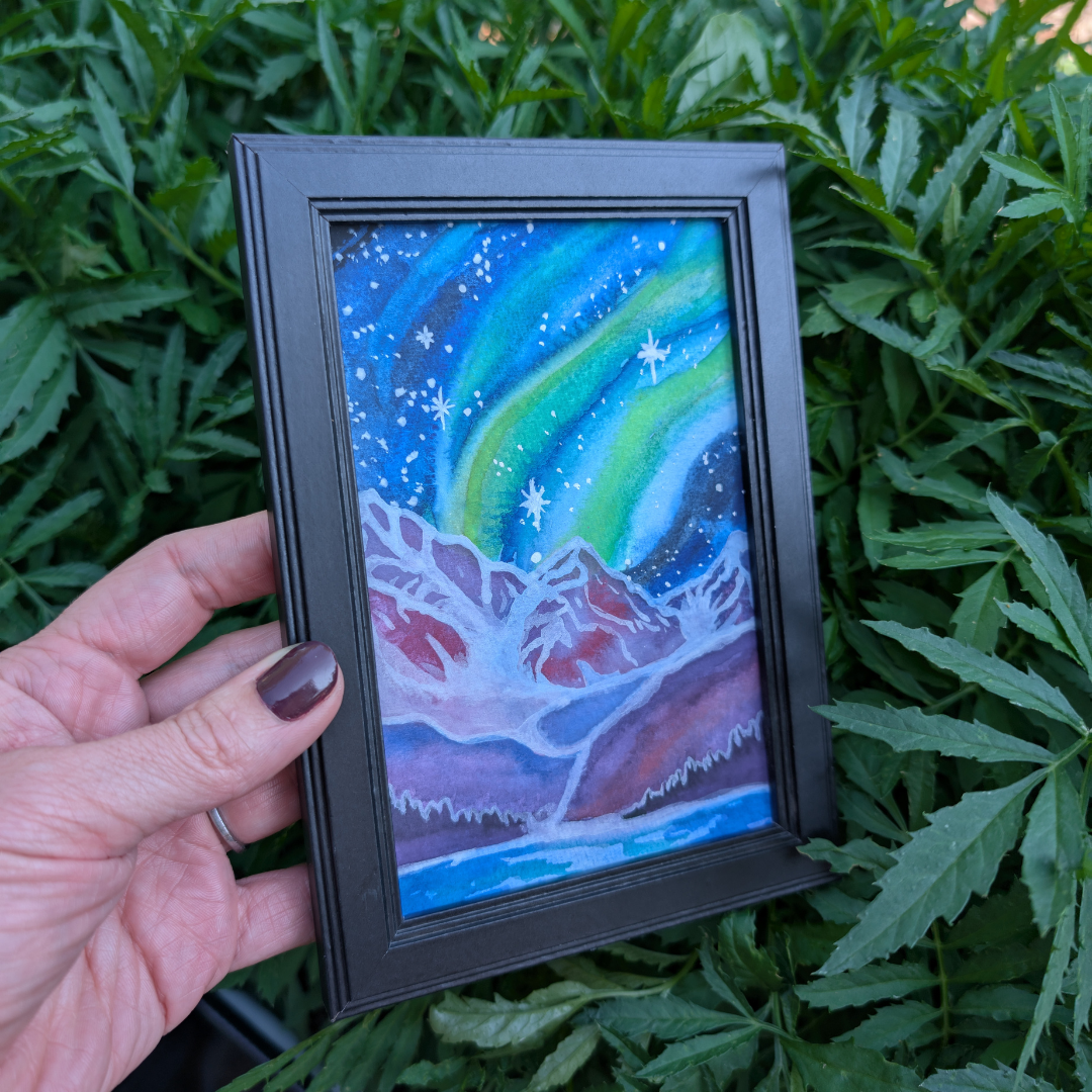 A framed painting of the northern lights over snowy mountains and a river, held in front of green foliage.