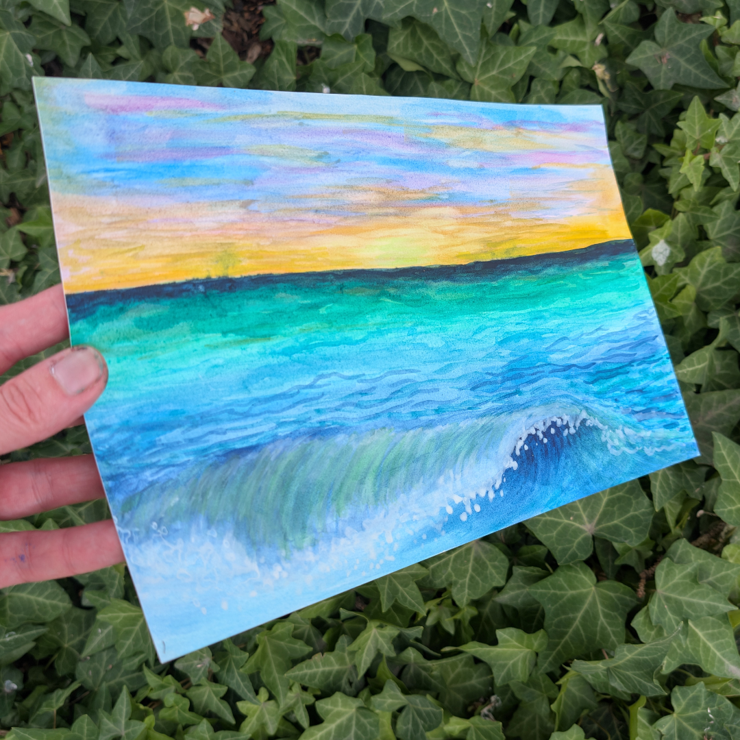 A hand holding a watercolor painting of a seascape with a sunset sky over the ocean, surrounded by green ivy leaves.