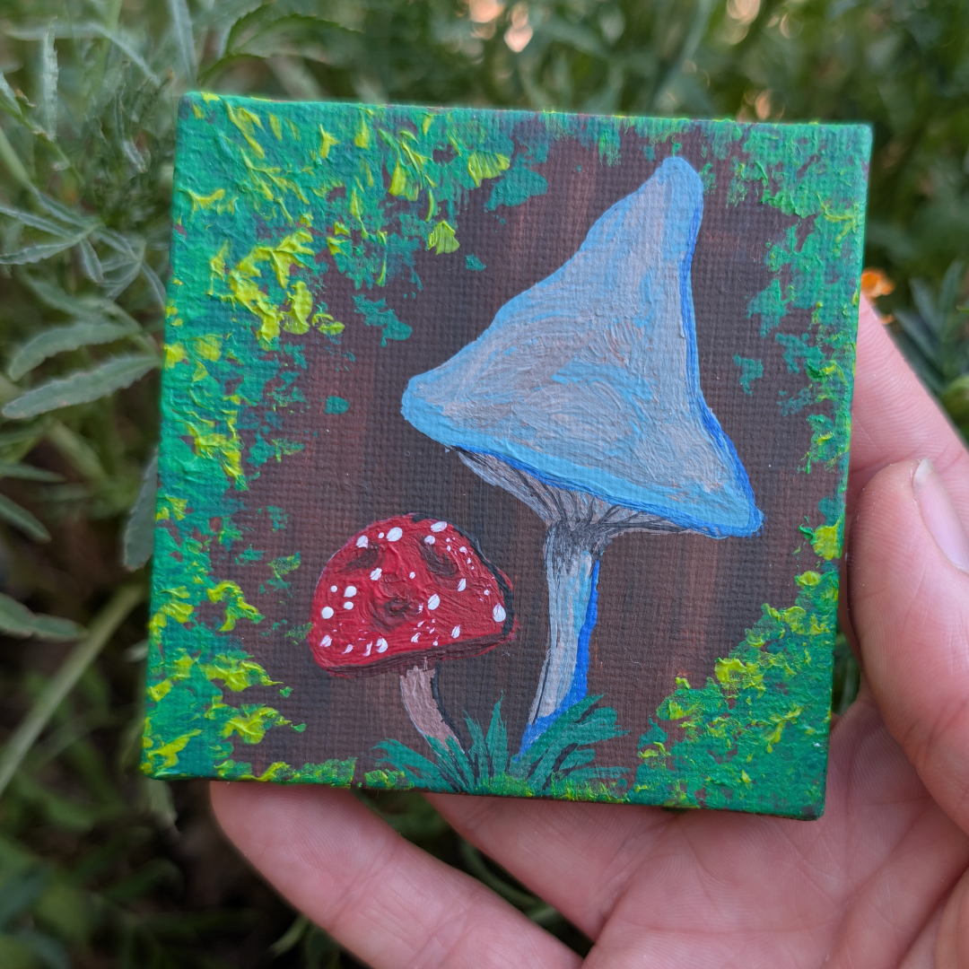 Small painted canvas featuring two mushrooms, a large blue one and a smaller red one with white spots, surrounded by green foliage.