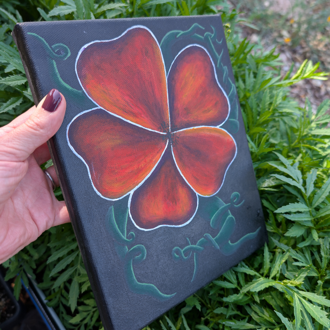 A canvas with a painting of a four-leaf clover in red, orange, and yellow shades outlined in white, surrounded by green decorative swirls, held by a person with burgundy nails among green foliage.
