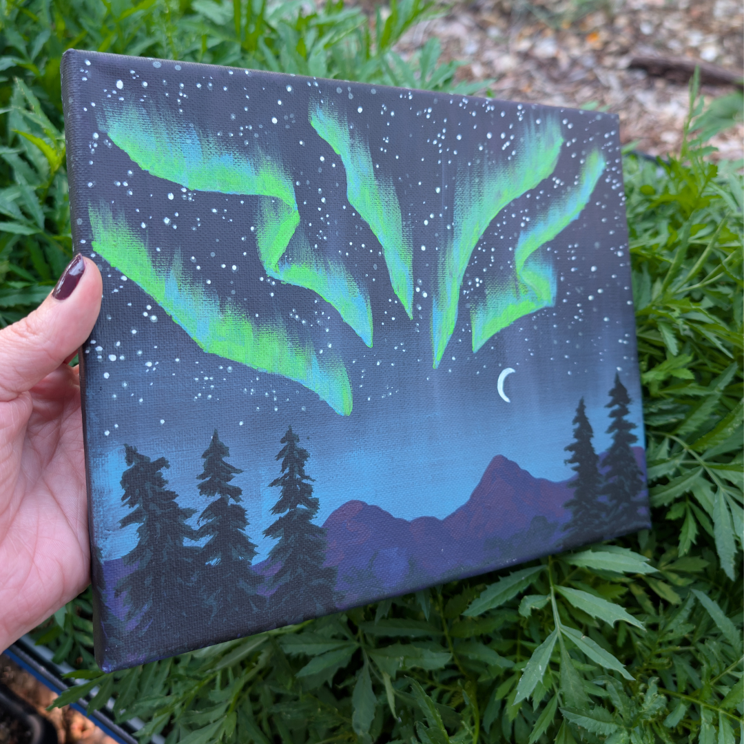A hand holding a painting of the night sky with the northern lights, stars, a crescent moon, mountains, and pine trees in the foreground.