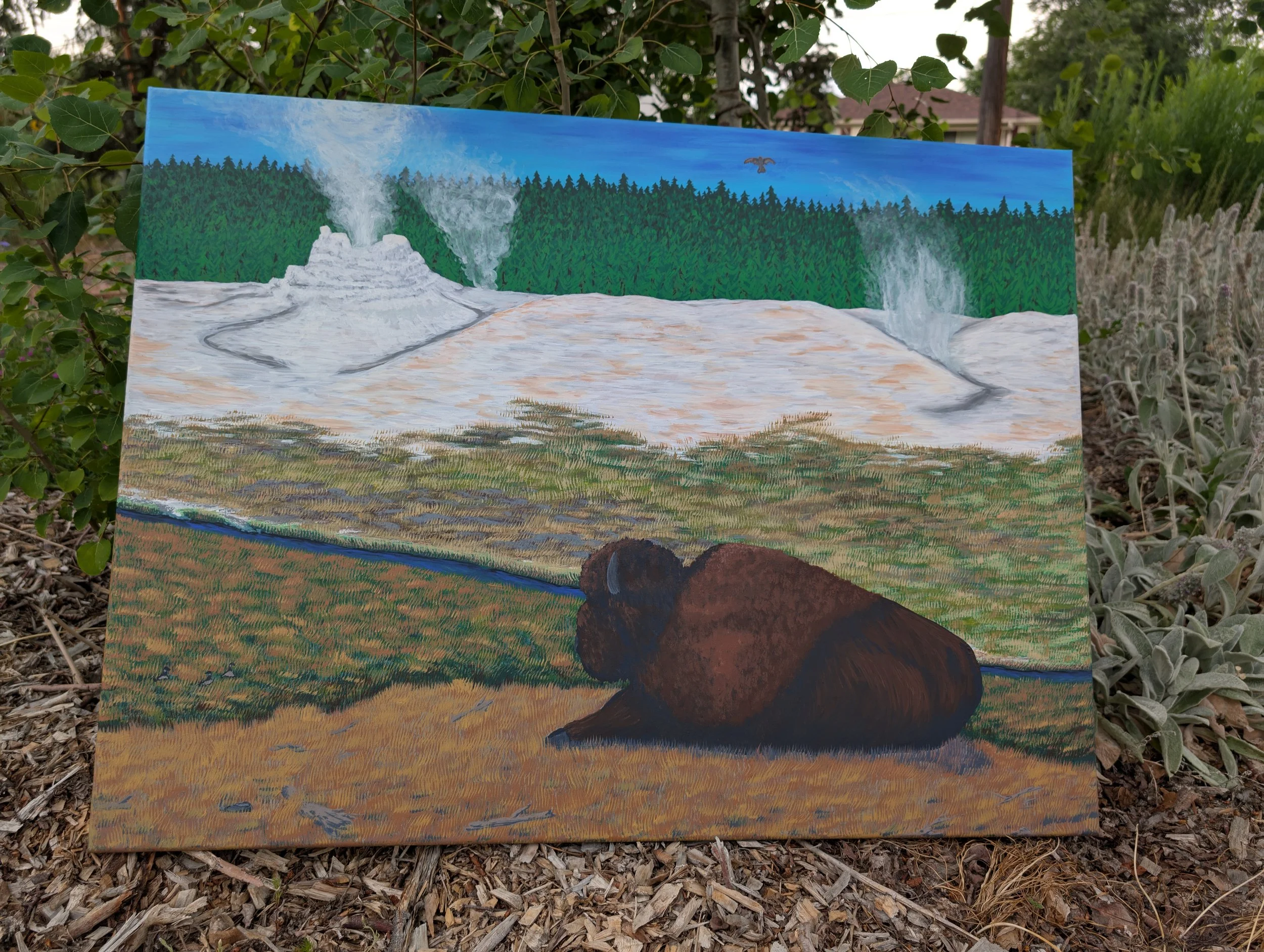 A painting of a brown bear lying on the ground in front of a landscape with waterfalls, a forest, and a large body of water, displayed outdoors among plants and mulch.