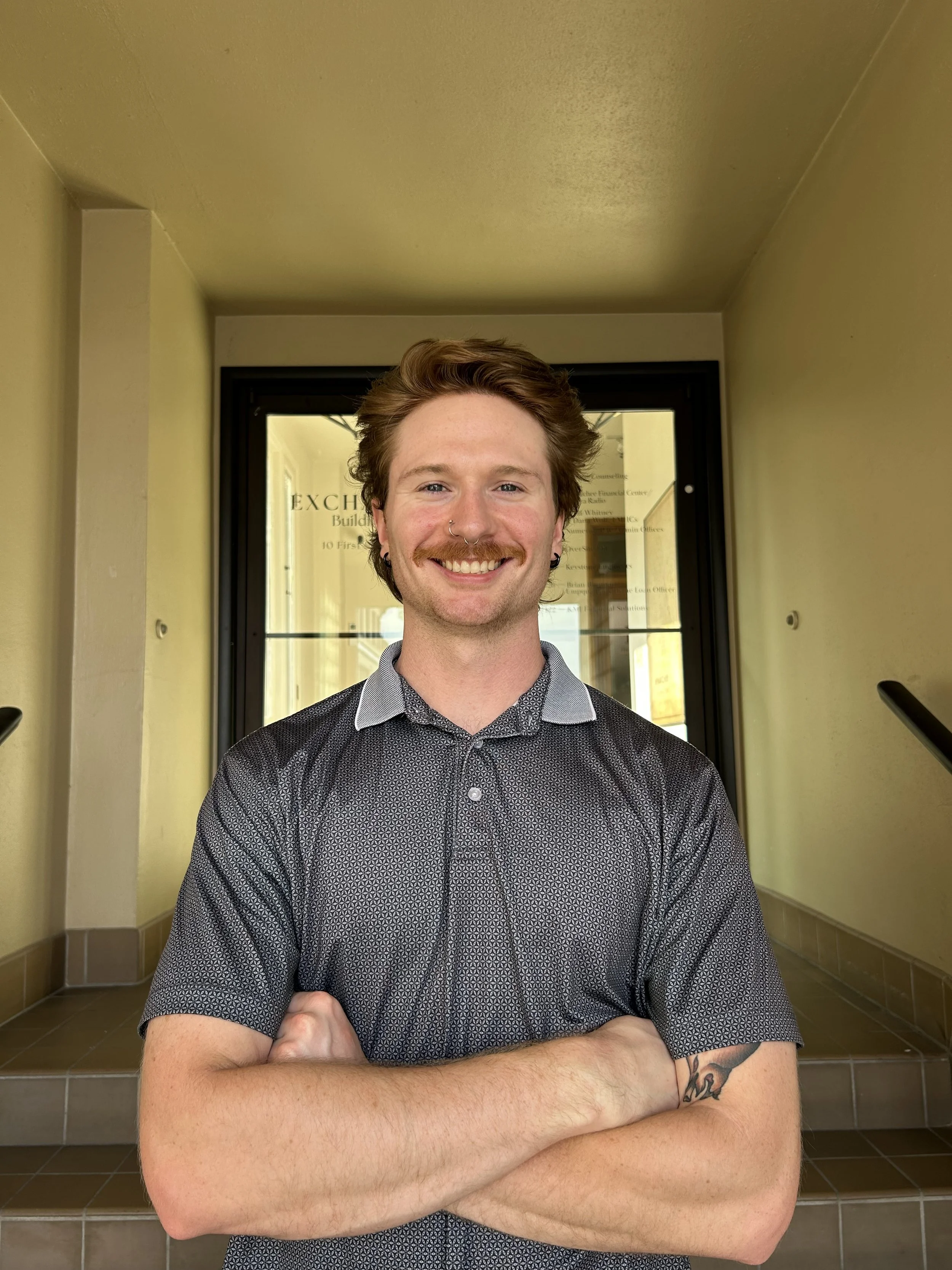 A young man with auburn hair, wearing a gray patterned shirt, standing with arms crossed in a building corridor with glass doors in the background.