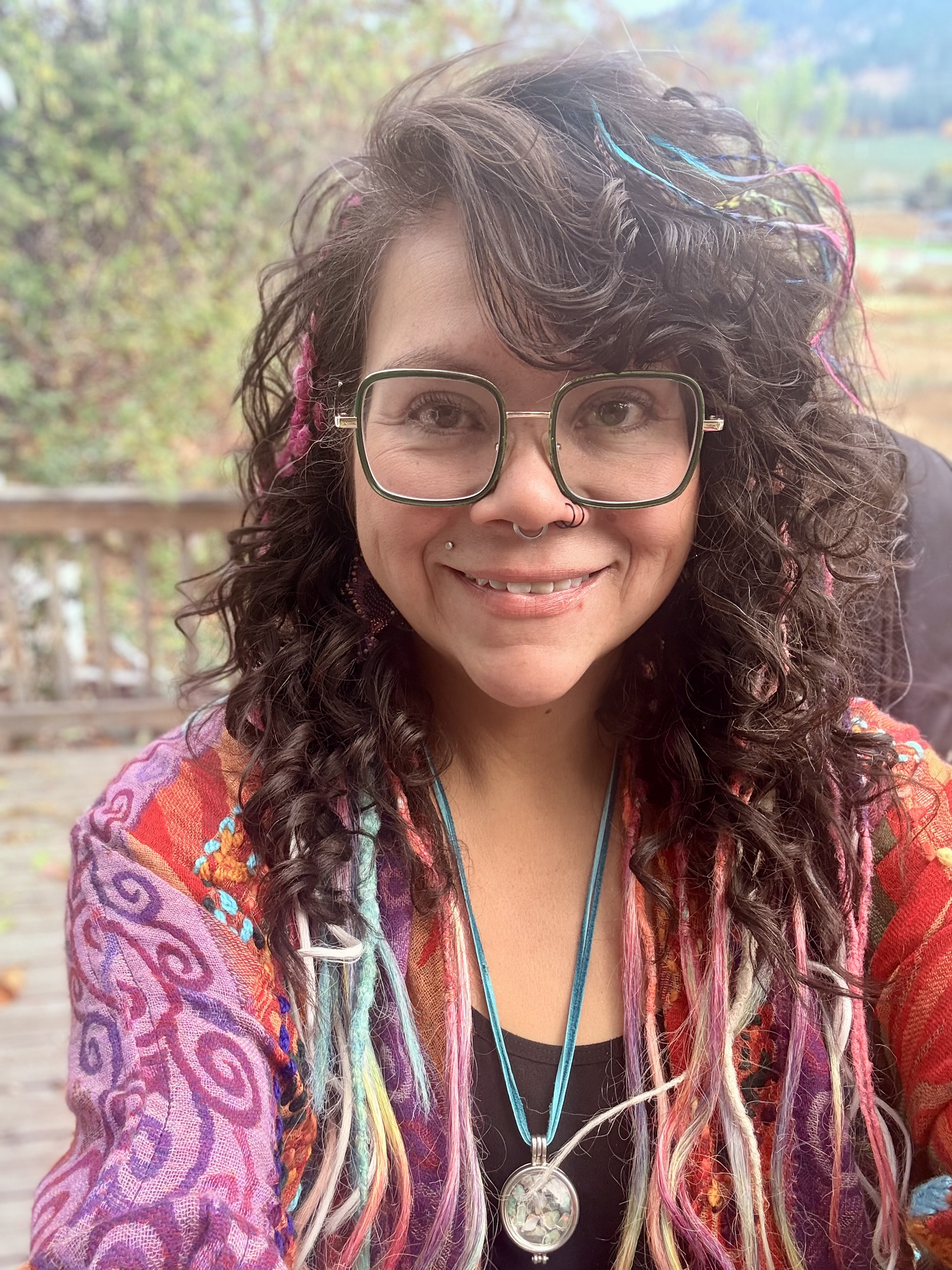 Close-up of a smiling woman with glasses, colorful dreadlocks, and a vibrant patterned jacket outdoors with autumn foliage in the background.