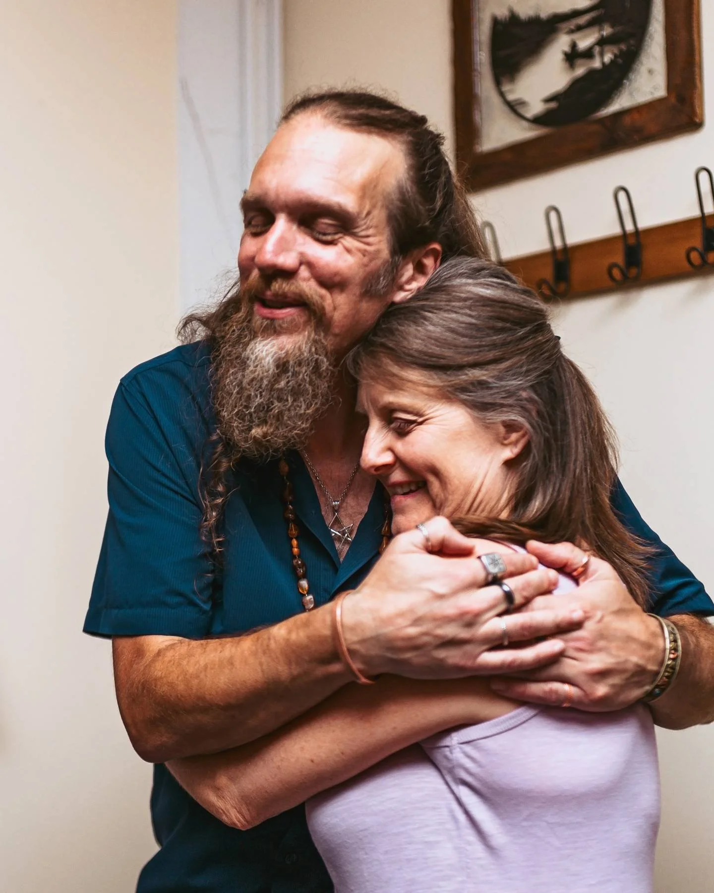 A man with a beard and long hair embracing a woman with brown hair in a hug, both smiling with eyes closed, indoors against a plain wall with a framed painting and hooks in the background.