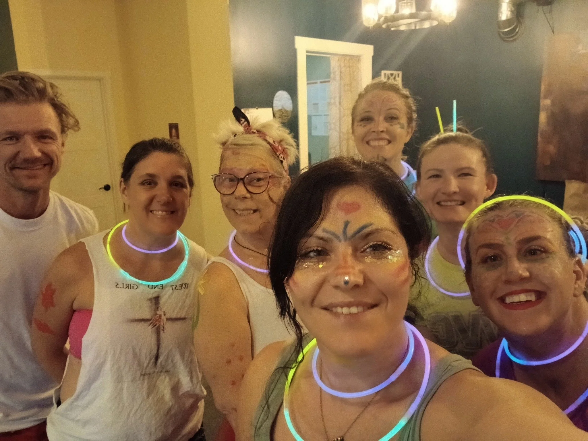A group of adults and children with face paint and glow-in-the-dark necklaces, smiling for a group photo in an indoor setting.