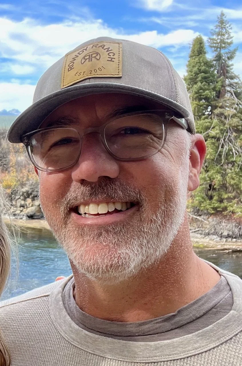 Smiling man wearing a blue shirt standing outdoors with a blurred background.