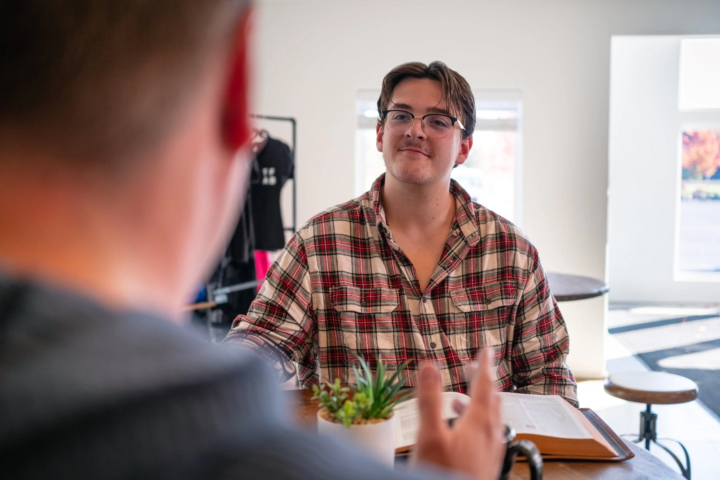 A man with glasses and a plaid shirt is sitting at a table, looking at another person who is gesturing with their hand. The background includes bright windows, a small plant, and some clothing on display.