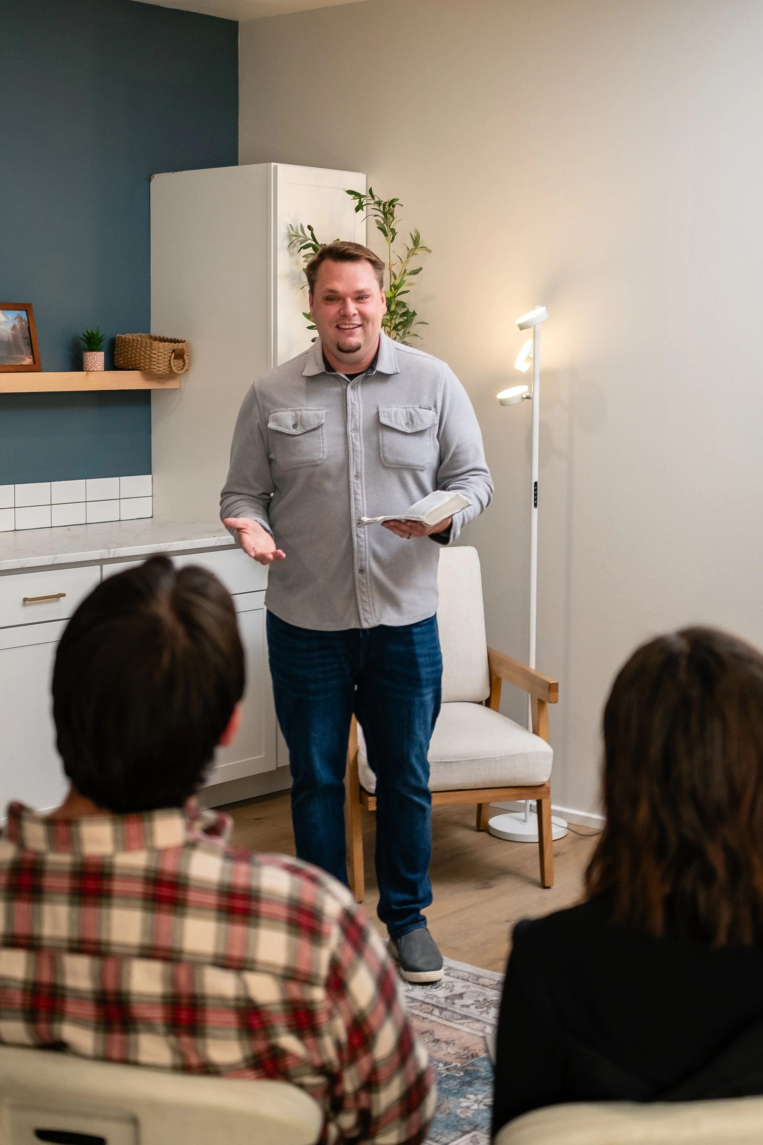 A man is speaking and holding a book in a living room, with two people sitting and listening to him.