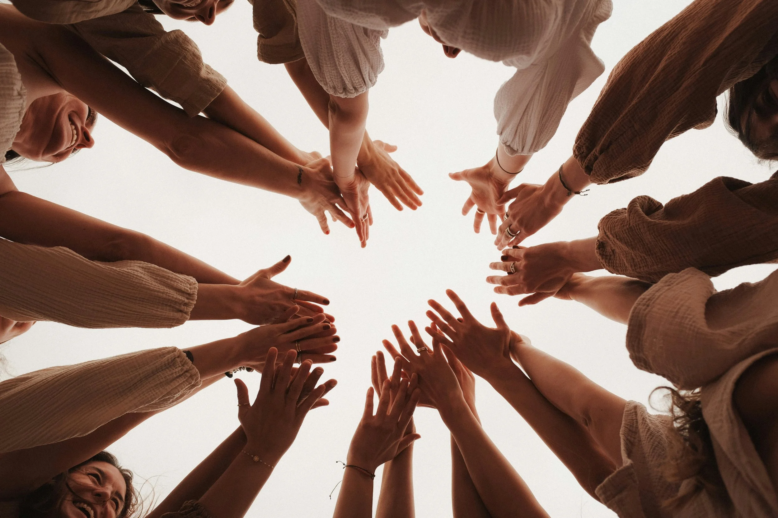 A group of people reaching their hands toward the center in a circle, seen from below against a plain background.