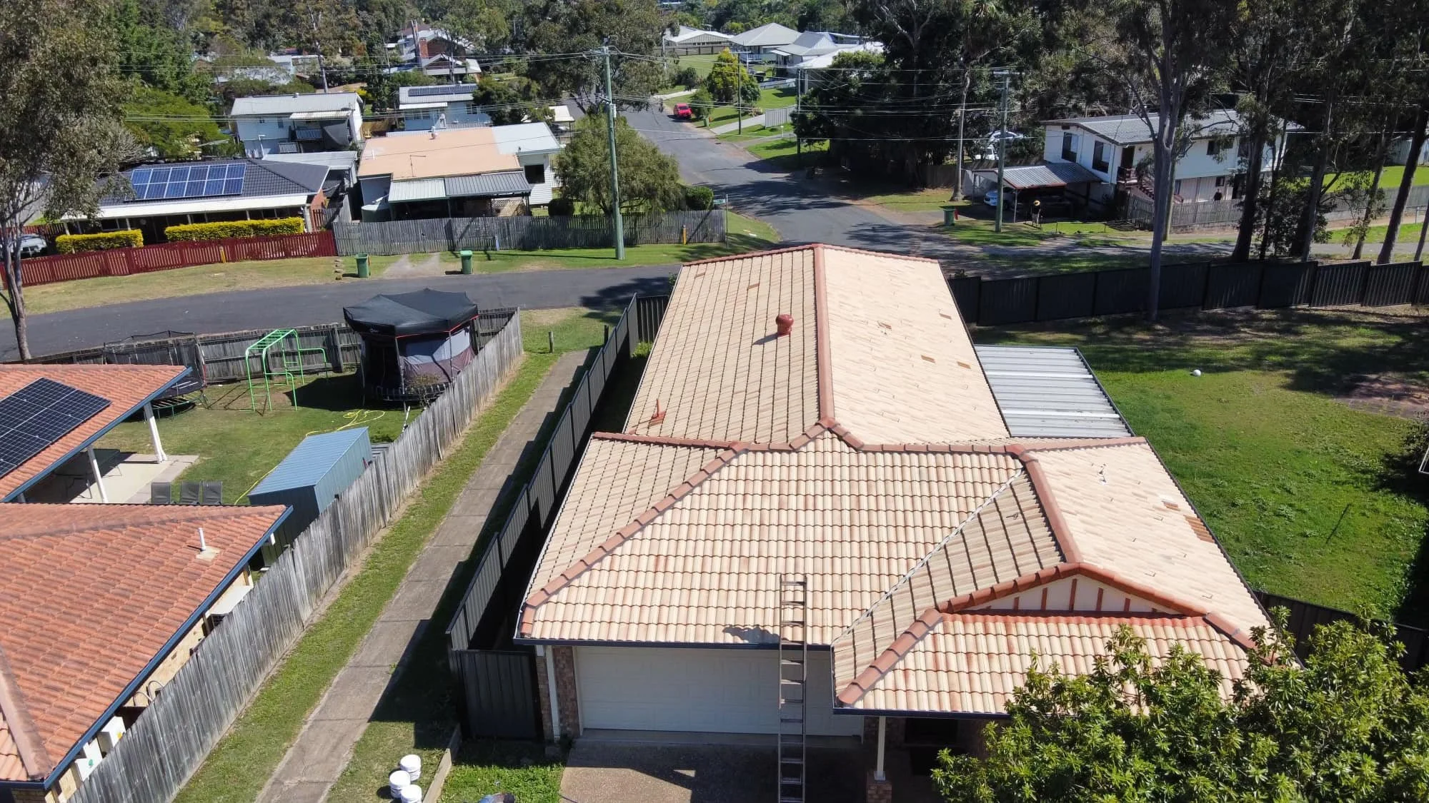 Aerial view of a residential neighborhood showing a house with a tiled roof, fenced backyard with a trampoline, garden shed, and neighboring houses with solar panels, on a sunny day.