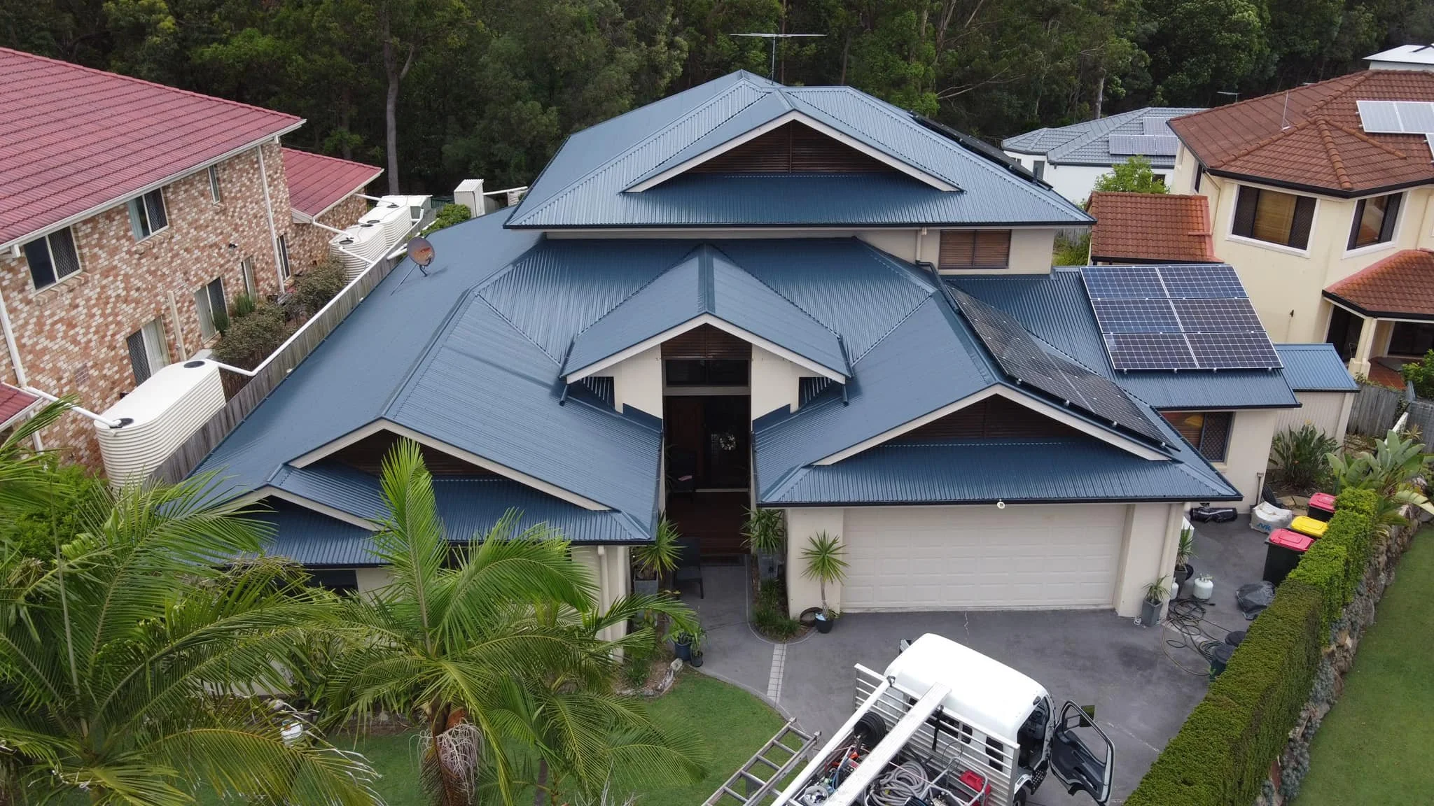 An aerial view of a modern house with a blue metal roof, solar panels, surrounding gardens, and neighboring houses under a green canopy.