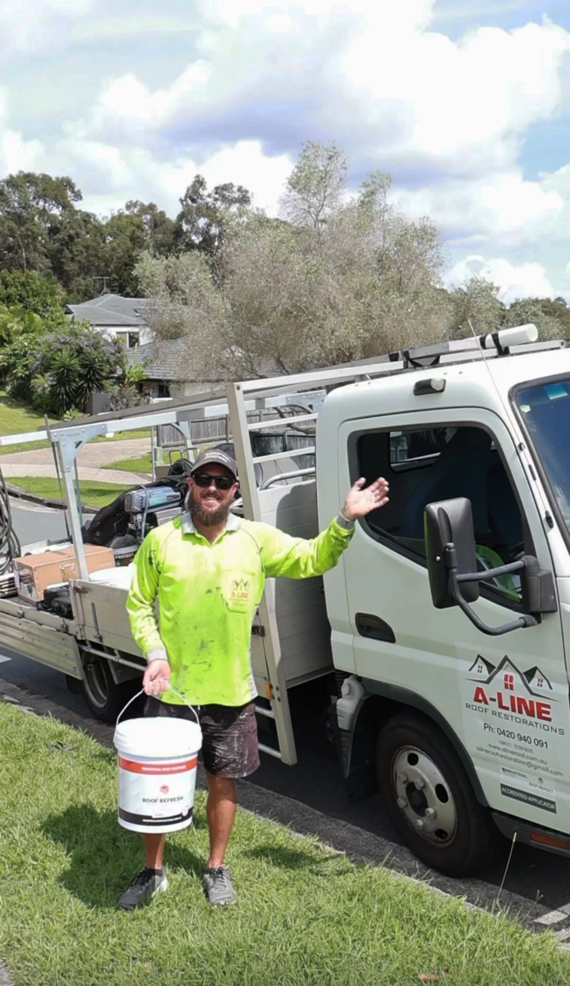 A man in a fluorescent yellow work shirt holding a bucket, standing next to a white truck labeled 'A-LINE Roof Restorations'. The man is smiling and gesturing with his hand. The truck is parked on a grass verge next to a residential street, with trees and houses in the background.