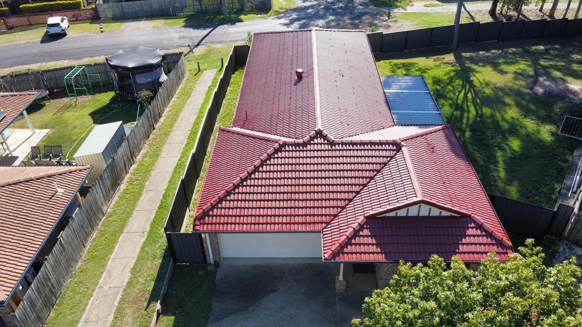 Aerial view of a house with a red tiled roof, a fenced backyard with a trampoline and a shed, and a sidewalk next to the house.