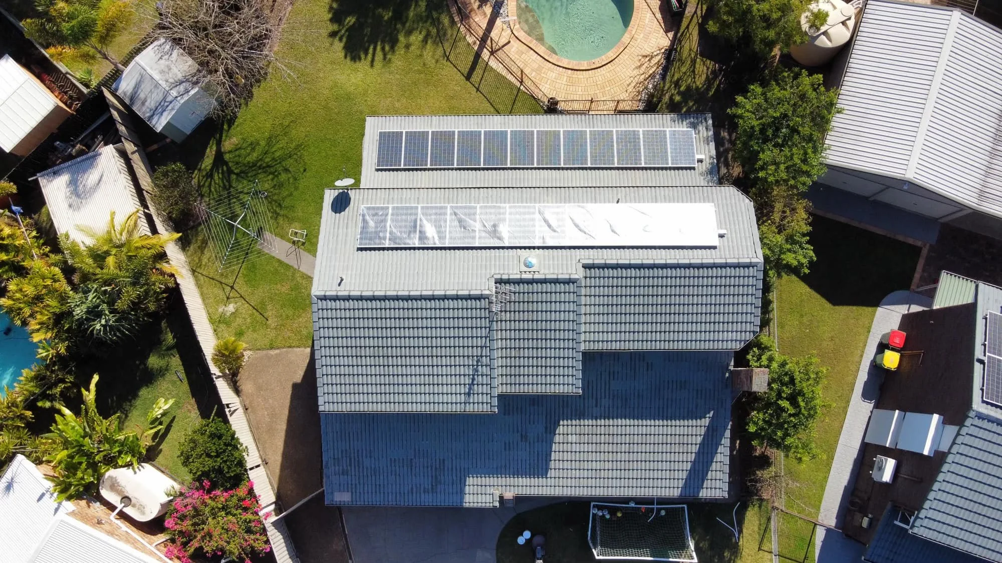 Aerial view of residential backyard featuring a house with solar panels on the roof, a trampoline, a swimming pool, a grassy area with a clothesline, and surrounding greenery.
