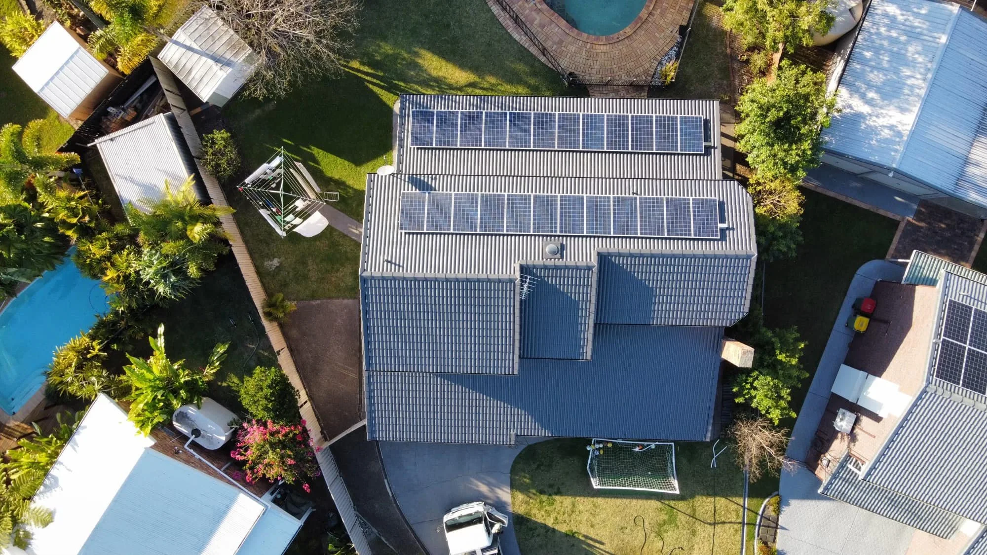 Aerial view of a residential backyard with solar panels on roofs, a swimming pool, a lawn, trees, a small basketball hoop, a parked car, and various outdoor structures.