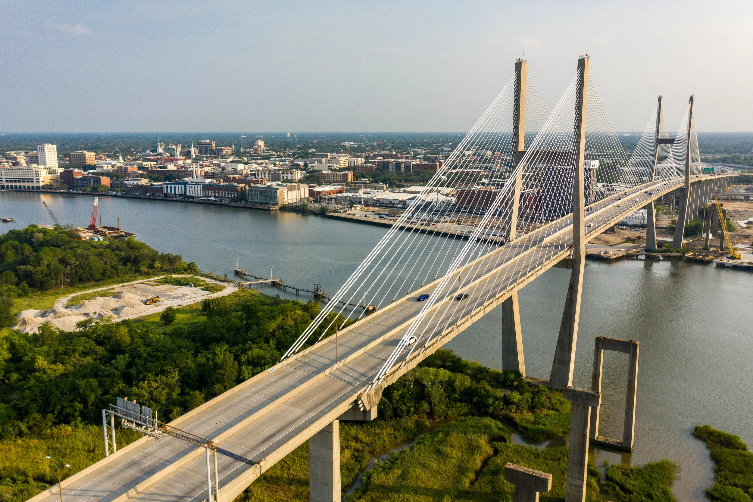 Aerial view of a cityscape featuring a large cable-stayed bridge over a river, with buildings, green areas, and a distant skyline under a partly cloudy sky.