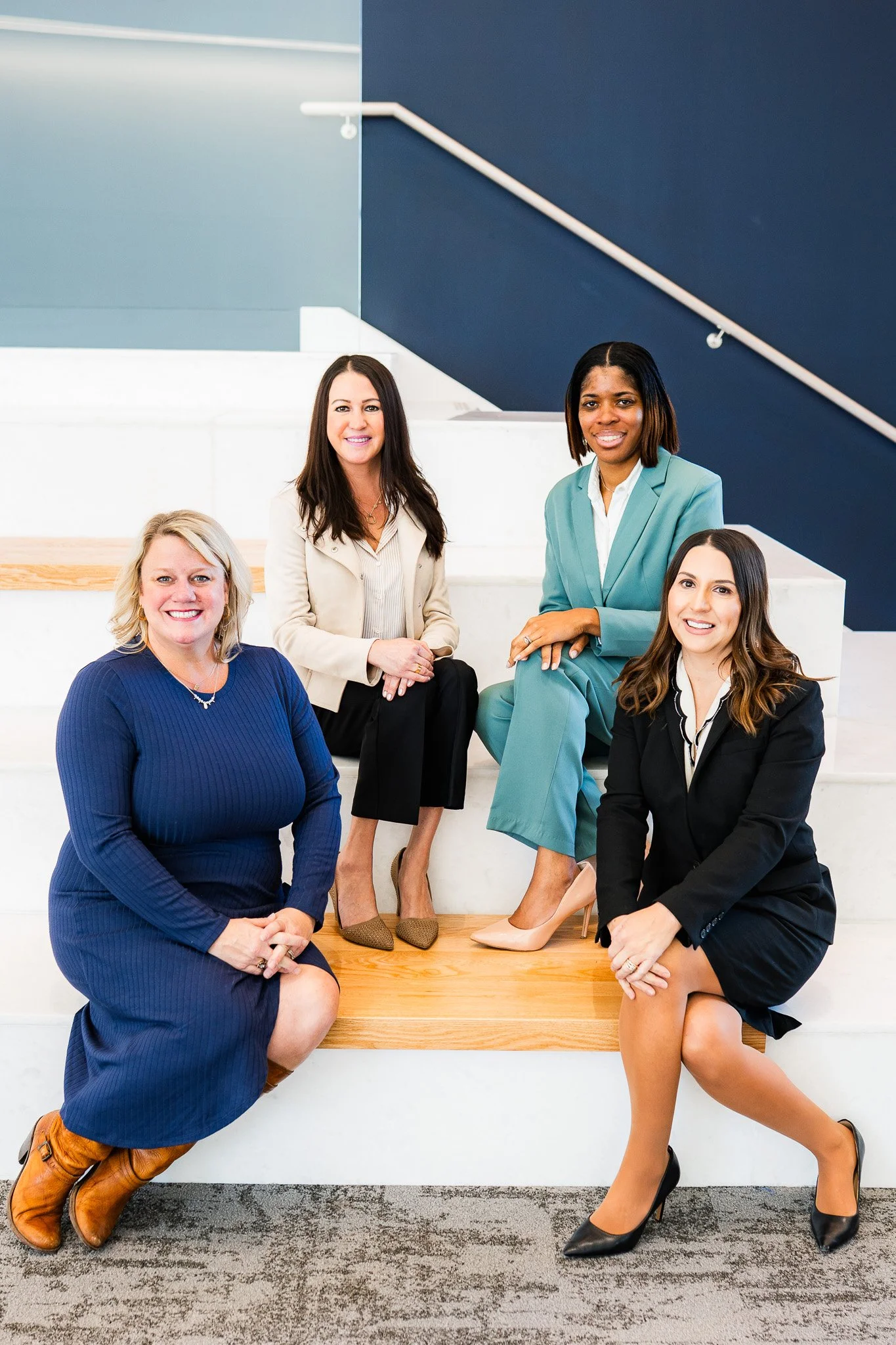Group of five women in business attire sitting and standing on white stairs in an office building with navy blue and light blue walls.