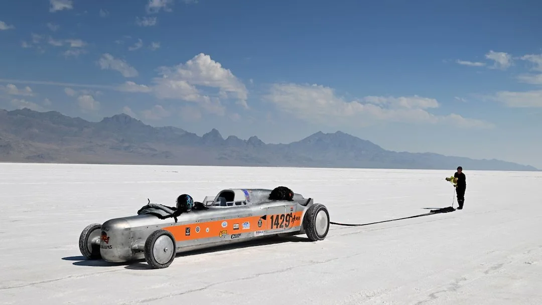 A streamlined, silver land speed racing vehicle with orange accents and various sponsor logos, connected by a tether to a person standing on a vast, flat salt flat with mountains in the background under a partly cloudy sky.