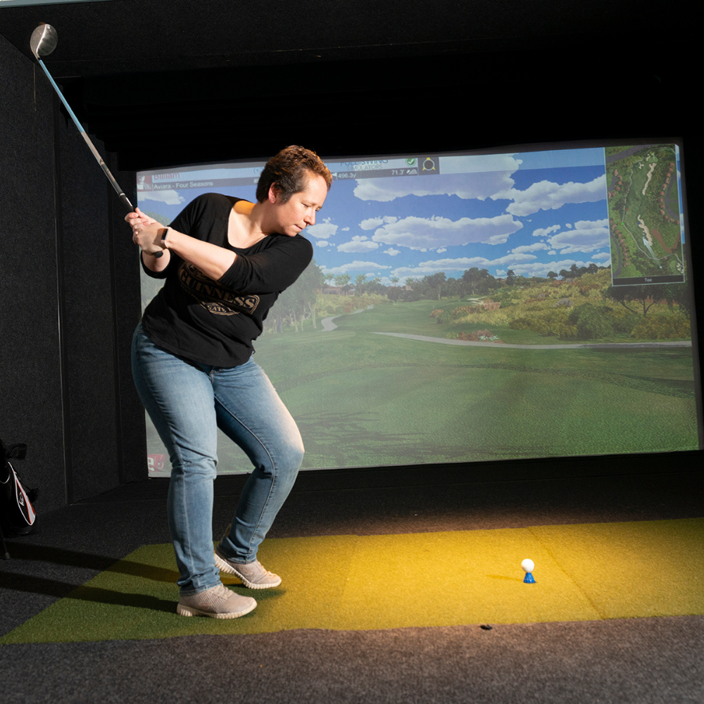 Group of six people, four women and two men, sitting and socializing in a room with a large indoor golf simulator screen. One man is swinging a golf club, hitting a ball towards the screen. The table has snacks and drinks, including a plate of appetizers, a cup of cocktail, and a glass of wine.