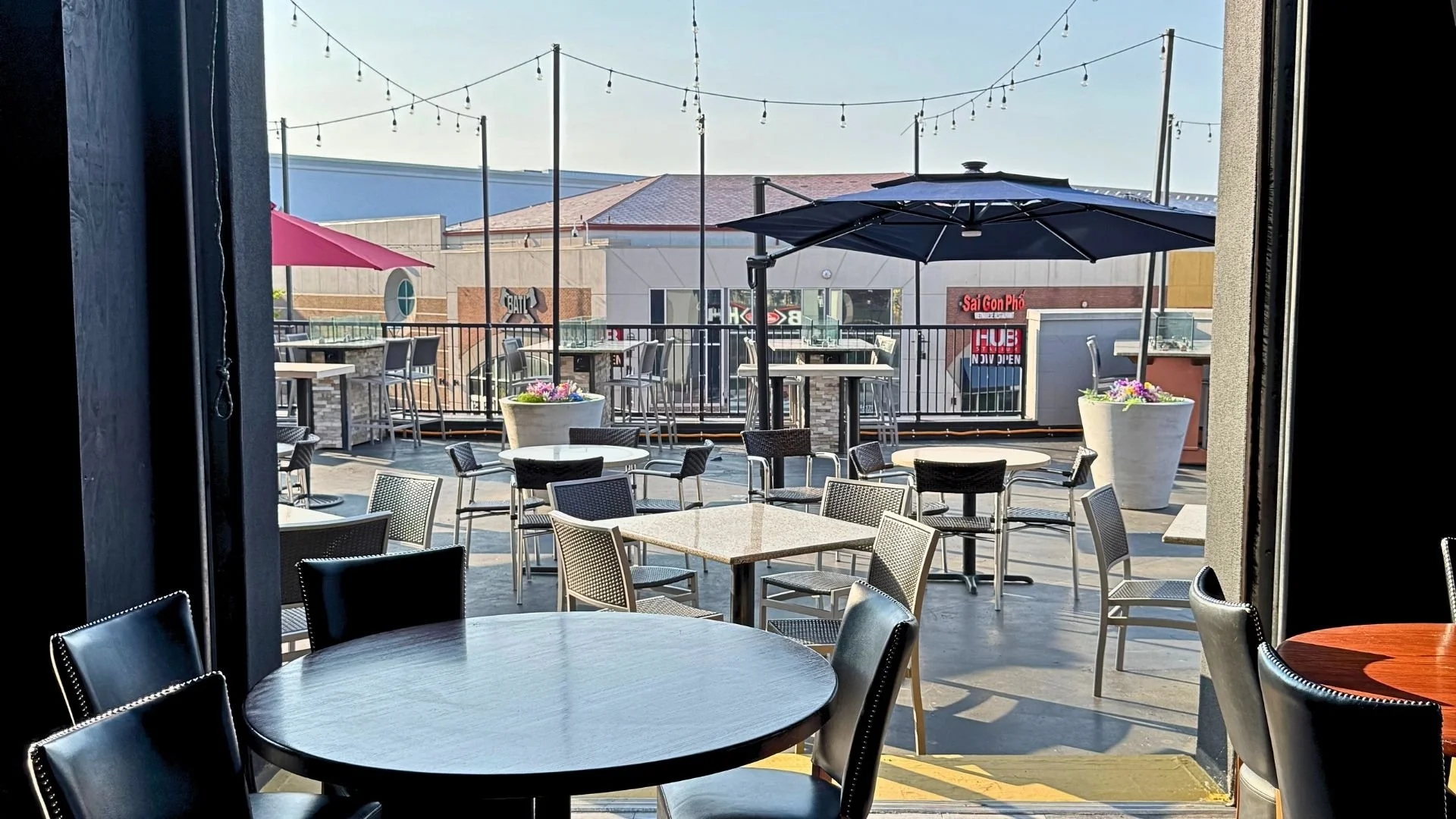 Outdoor patio of a restaurant with empty tables and chairs, large potted plants, umbrellas, string lights, and a nearby shopping center building.