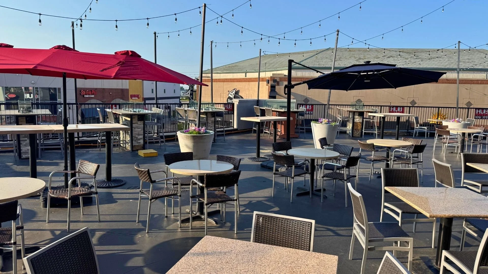 An outdoor restaurant patio with tables, chairs, umbrellas, and string lights overhead. There are large planters with flowers and a building in the background.