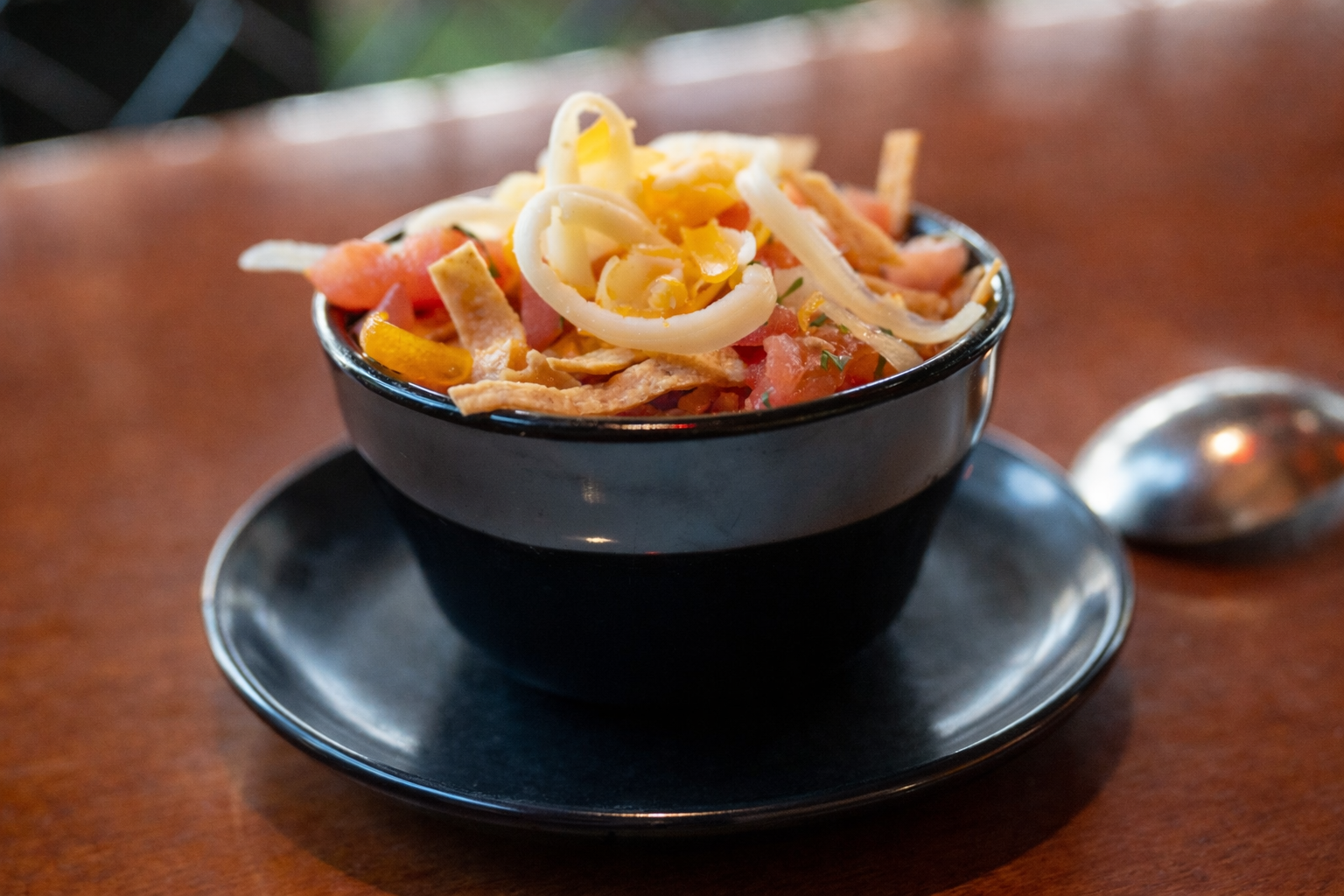 A bowl of taco salad topped with shredded cheddar cheese, sliced peppers, chopped tomatoes, and shredded chicken on a dark plate, with a spoon on a wooden table.