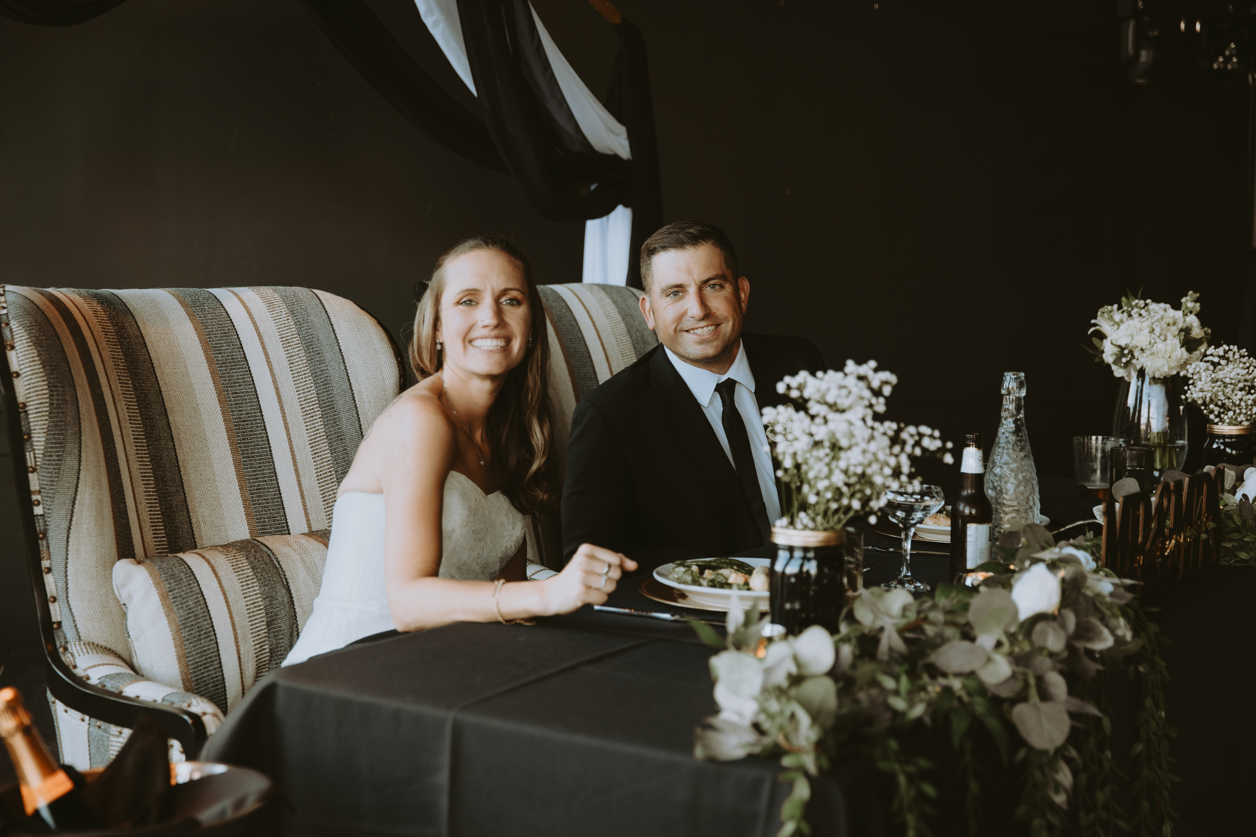 A smiling couple sitting at a decorated table during an event, with floral arrangements and drinks.