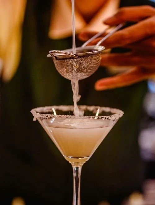 A martini glass with a salted rim holding a creamy cocktail, and a tea strainer with a wire handle releasing liquid into the drink.