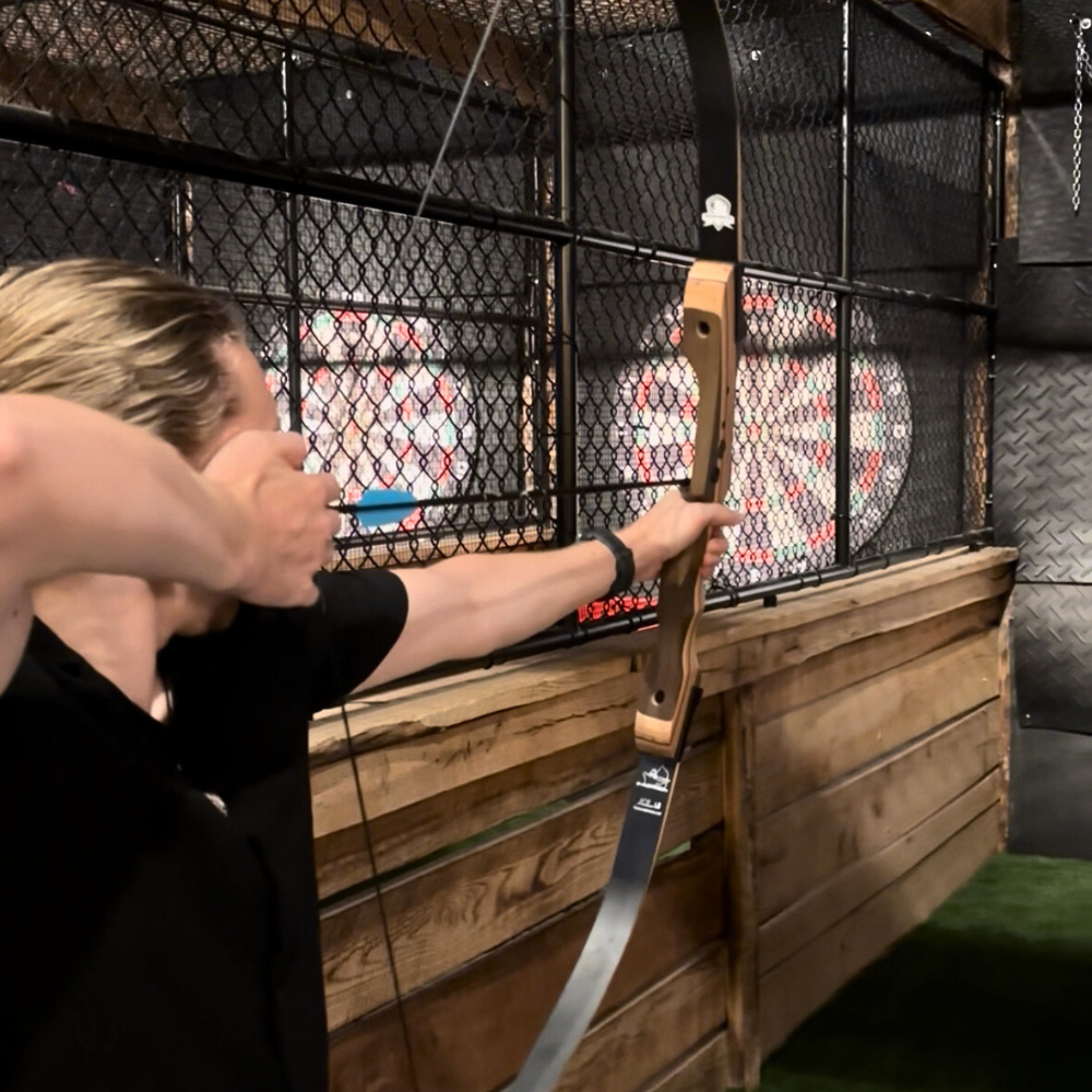 Person aiming a bow and arrow at an archery target inside an indoor range with a wooden and metal structure.