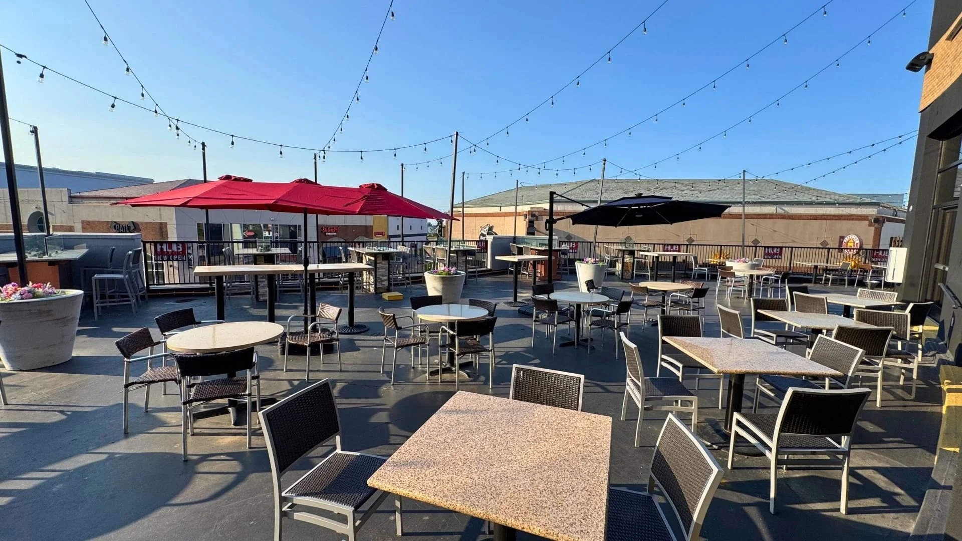 Empty outdoor patio with tables, chairs, large umbrellas in red and black, string lights overhead, and potted plants on a sunny day.