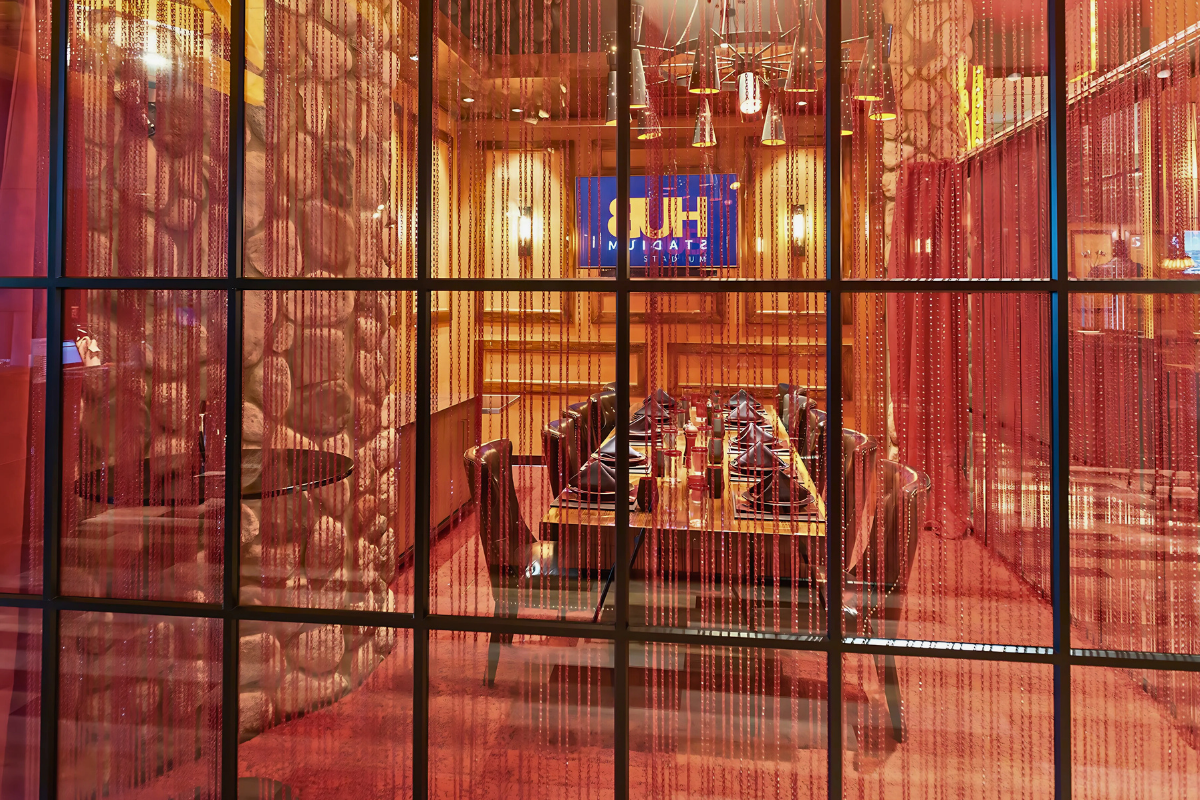 View of a modern restaurant interior through glass windows, with a long dining table set with black napkins and red decor, and a screen displaying a logo in the background.