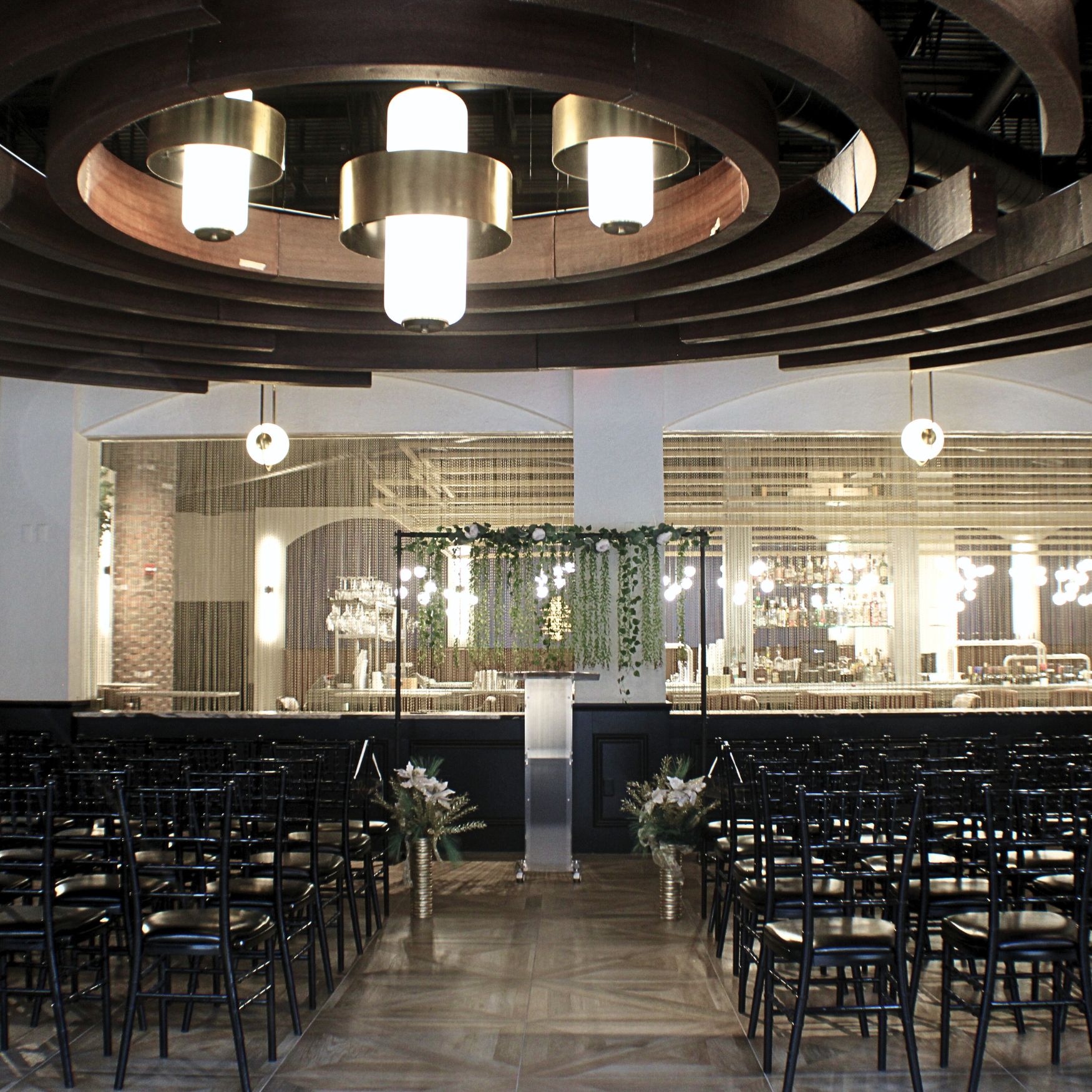 Elegant indoor wedding venue setup at The Hub Stadium Fountain Bleu Space with white chairs, a beautifully decorated altar, soft ceiling lighting, and a stylish bar area in the background. Perfect for weddings and special events in Novi, Michigan.