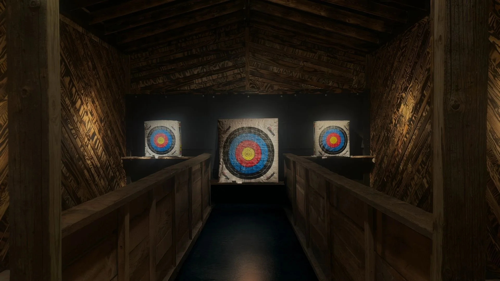 Indoor archery target range with three large circular targets on a black wall, framed by wooden walls and ceiling.