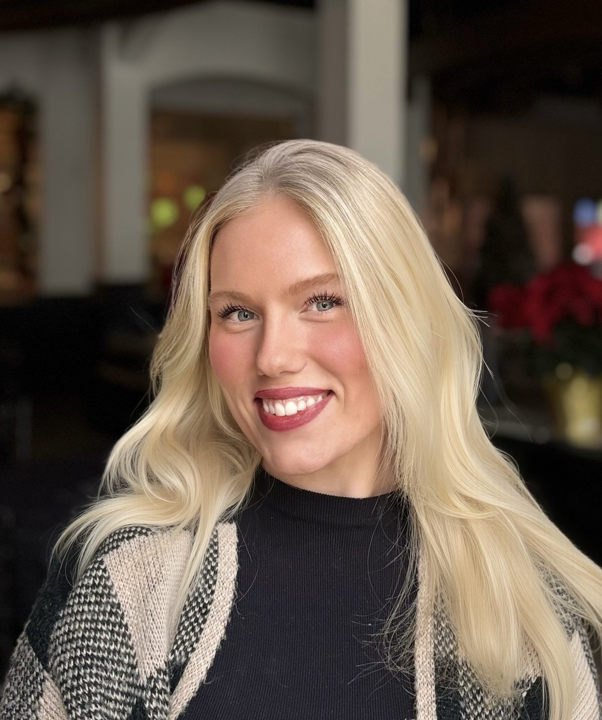 A smiling blonde woman with blue eyes, wearing a black top and a black-and-white patterned cardigan, in a warmly lit indoor setting.