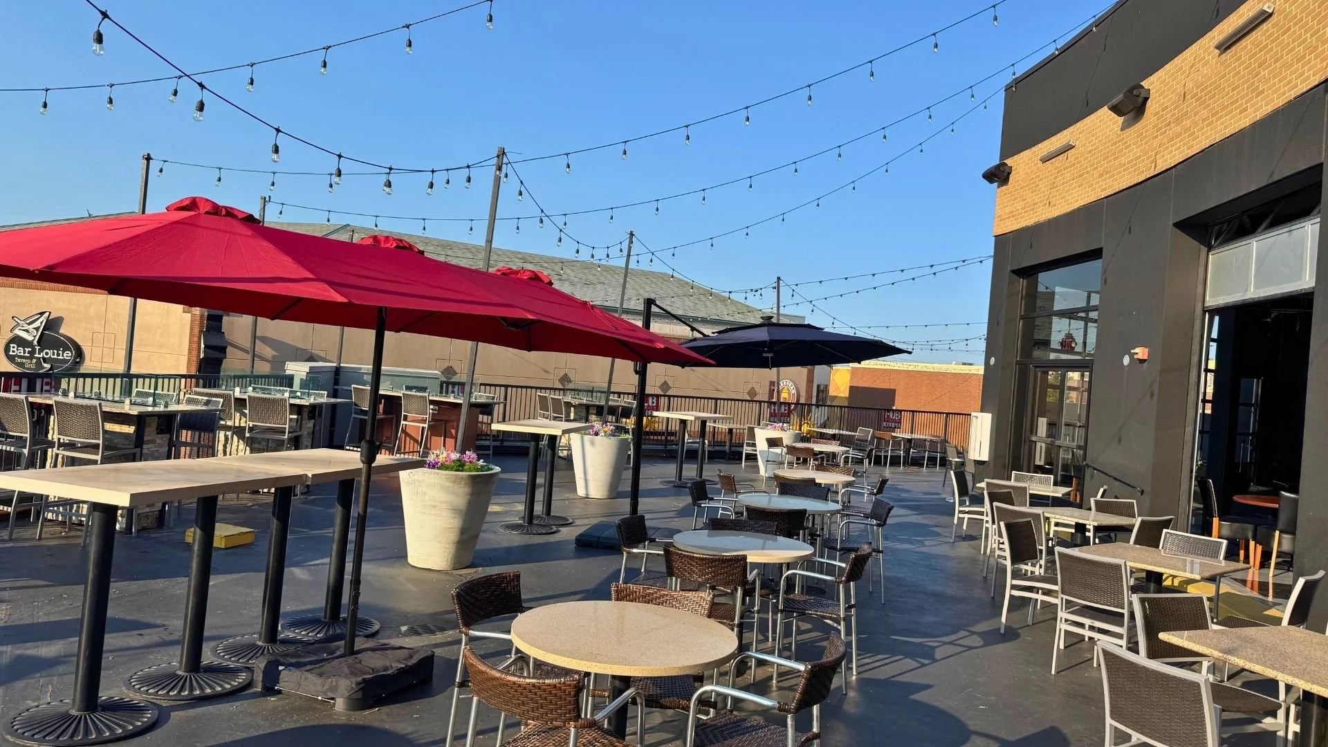 Empty outdoor restaurant patio with tables, chairs, large umbrellas, string lights, and a building wall with windows, under a clear blue sky.