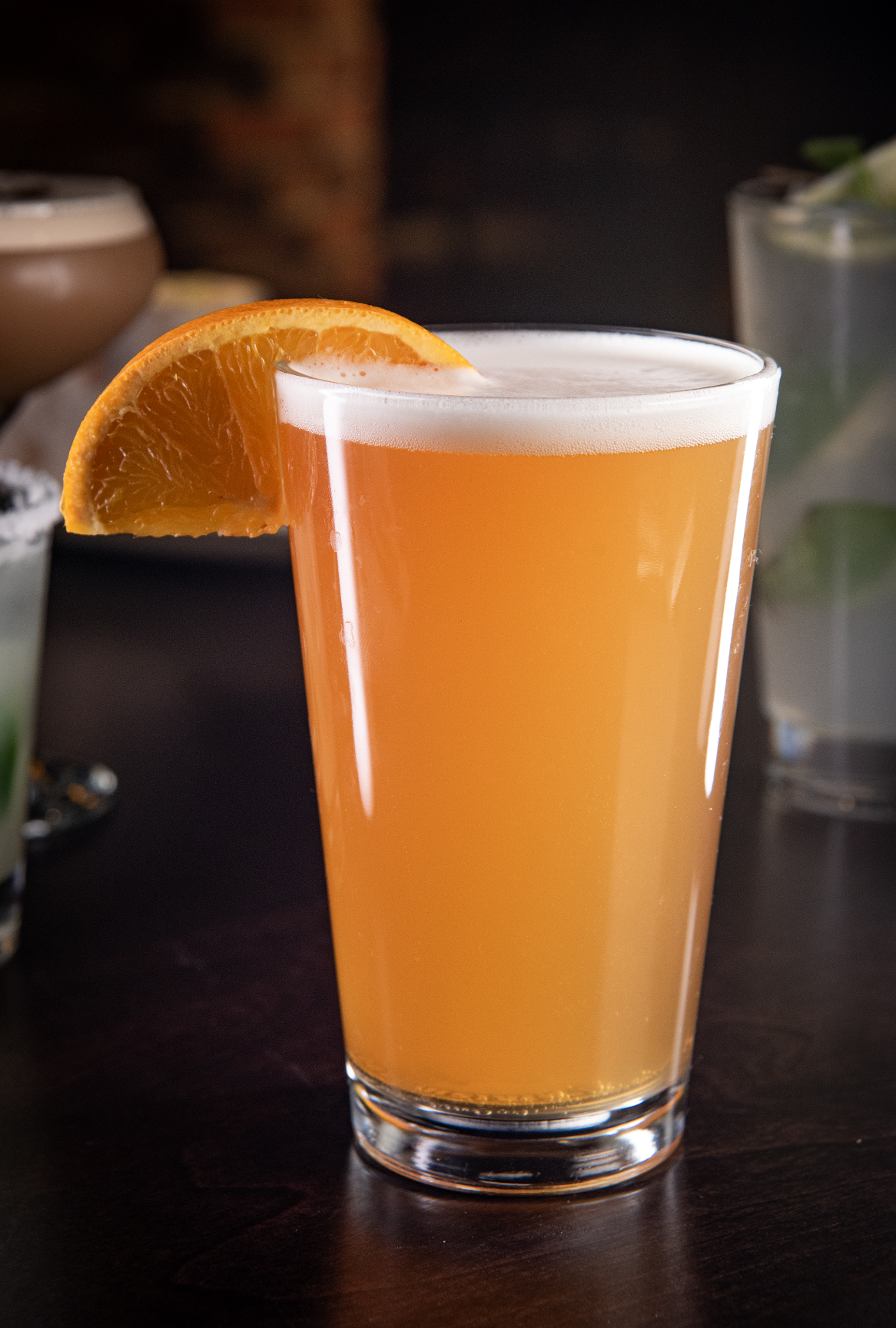 A tall glass of orange-colored drink garnished with a slice of orange on the rim, sitting on a dark surface.