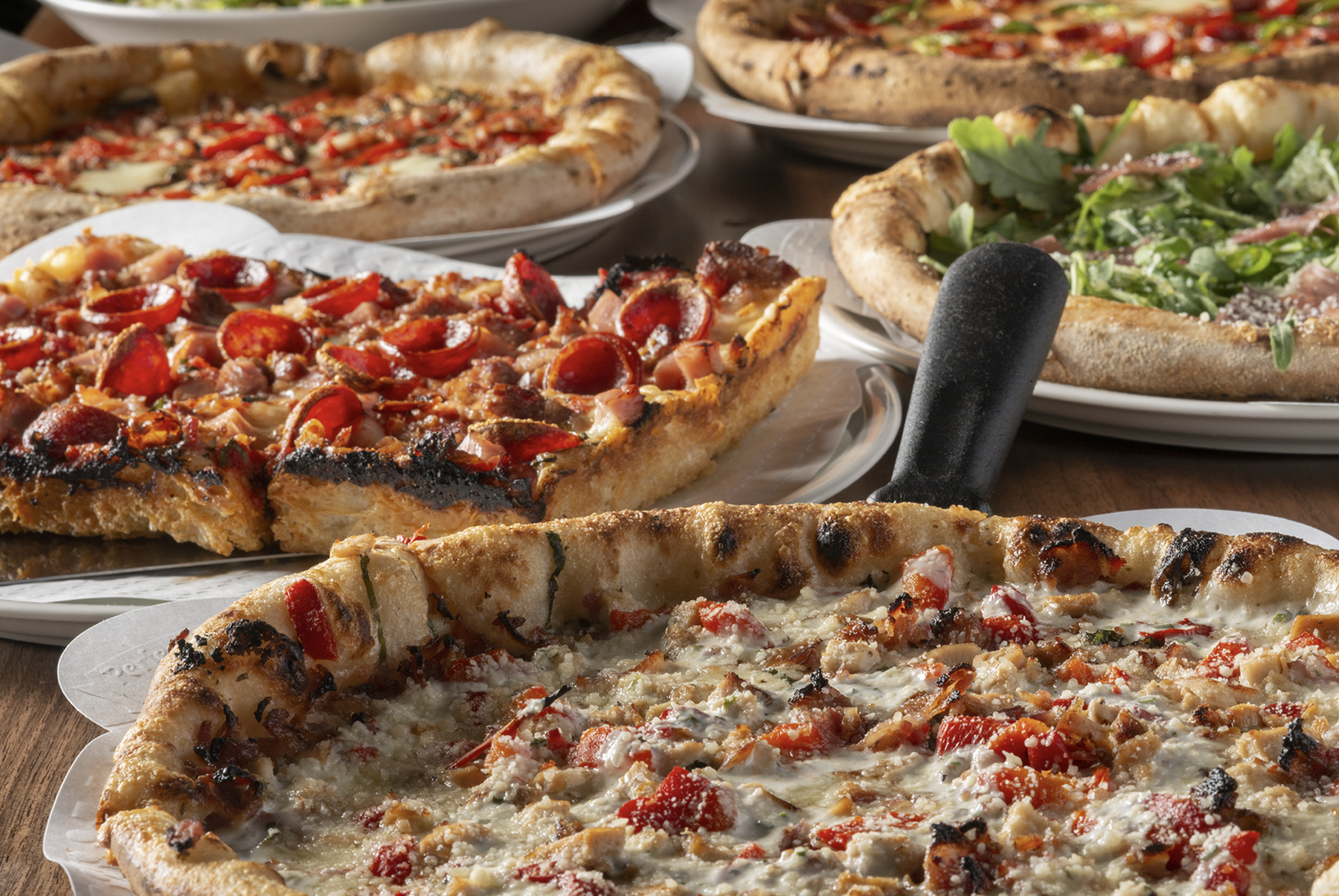 Several pizzas with different toppings, including vegetables, meats, and cheese, on a wooden table.