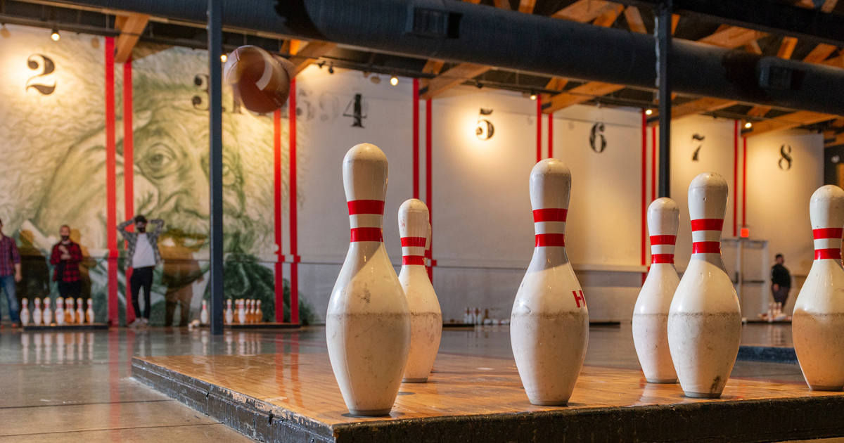 Indoor bowling alley with six large pins in the foreground, and a mural of Benjamin Franklin on the wall. There are people in the background near the pins and a mural. The alley has wooden floors and a high ceiling.