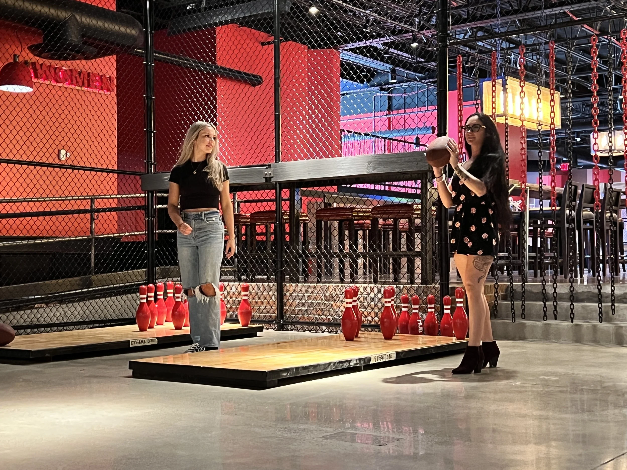 Two women at a bowling alley with red bowling pins, one holding a bowling ball and the other watching, in a colorful, modern setting.