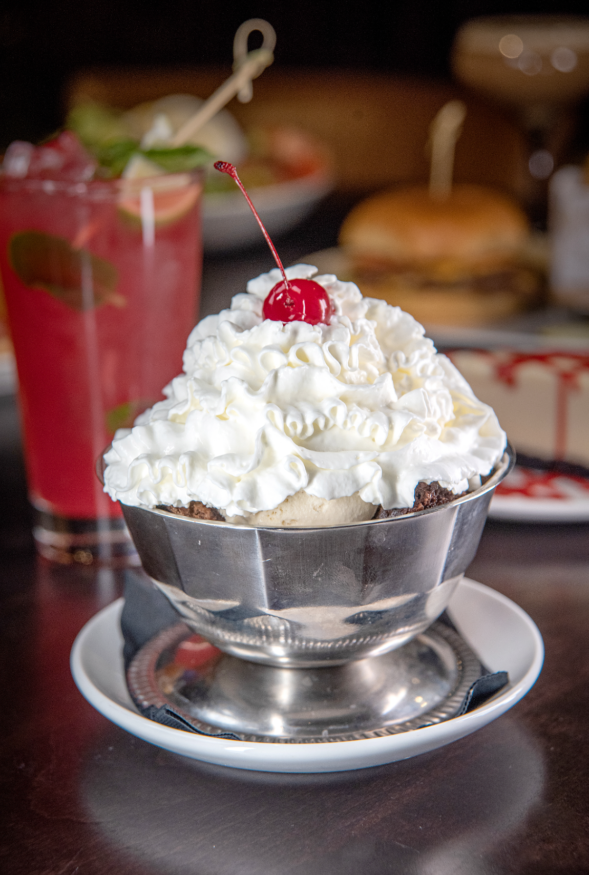 A dessert with whipped cream topped with a cherry in a stainless steel bowl on a white plate, with a pink drink and food in the background.